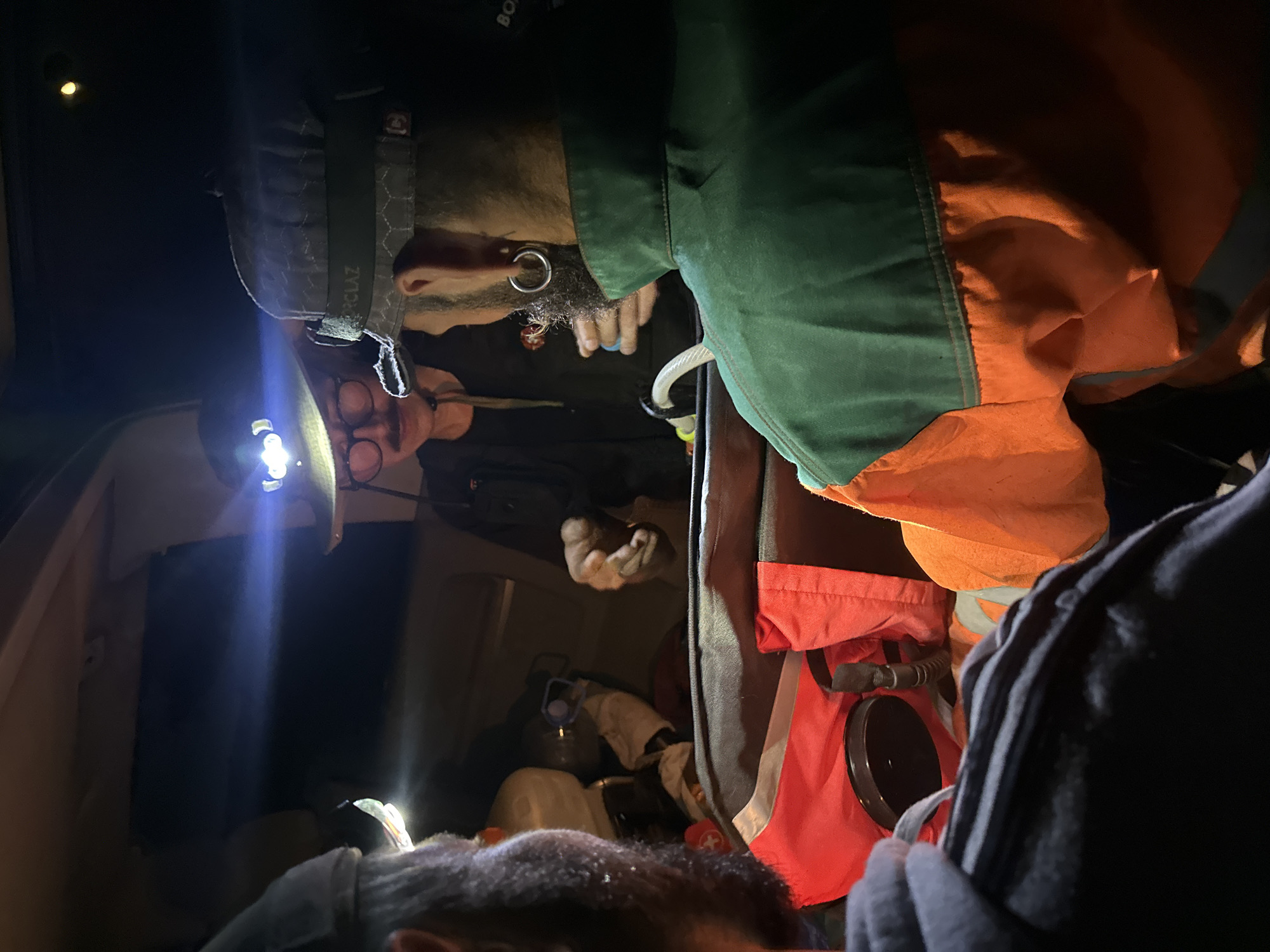 In Bijele Poljane, Montenegro, three individuals are inside a vehicle at night, illuminated by headlamps and interior lights. The scene is dark and appears to be contained within the rear compartment of the vehicle, possibly a van or SUV.
The person in the center, facing the camera, wears a wide-brimmed hat with a headlamp providing a bright beam of light. They have round glasses and a mustache. Their hands are visible, appearing to be interacting with something out of frame.
To the right of the central figure, another individual is seen from behind, wearing a cap with the logo "CLAZ" and a large hoop earring. They are wearing a green and orange vest. Their hands are also visible, suggesting an interaction or task.
To the left, the back of a third individual is visible, facing away from the camera, also wearing a cap. They have a beard and are partially obscured by darkness.
In the foreground, a large bag, possibly a duffel bag, is open, containing items such as a red vest or fabric and a black lid or cap. Other objects, including what appears to be a white gas canister with a blue handle, are visible within the vehicle's cargo area. The overall impression is one of a team engaged in some activity or preparation within a confined space at night.