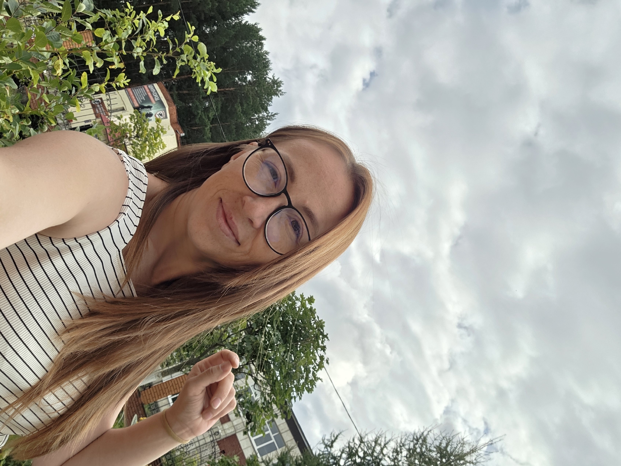 A blonde woman with glasses is taking a selfie outdoors. She has long, light brown hair and is wearing a white and black striped top. In the background, there are trees, a building, and a cloudy sky. The building in the background has Cyrillic writing that appears to say "ЦЕНТР-ГОРОД," which translates to "CITY-CENTER."