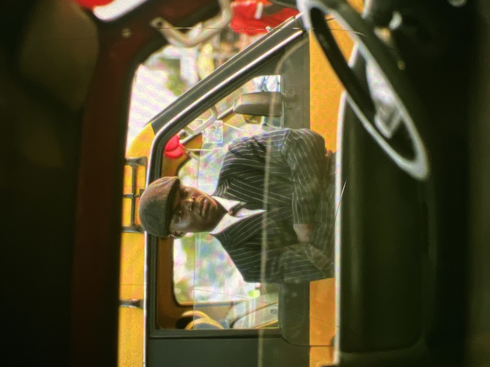 The image depicts a man wearing a pinstripe suit, a white shirt, a tie, and a flat cap, looking directly at the camera from inside a yellow vehicle, possibly a taxi, in Lagos, Nigeria. His arms are crossed. The vehicle's interior, including a steering wheel, is visible in the foreground. Reflections in the glass suggest an outdoor setting with other vehicles and people present. The overall lighting and image quality suggest it may be a candid shot or a still from a film. The atmosphere appears to be that of a bustling city environment.