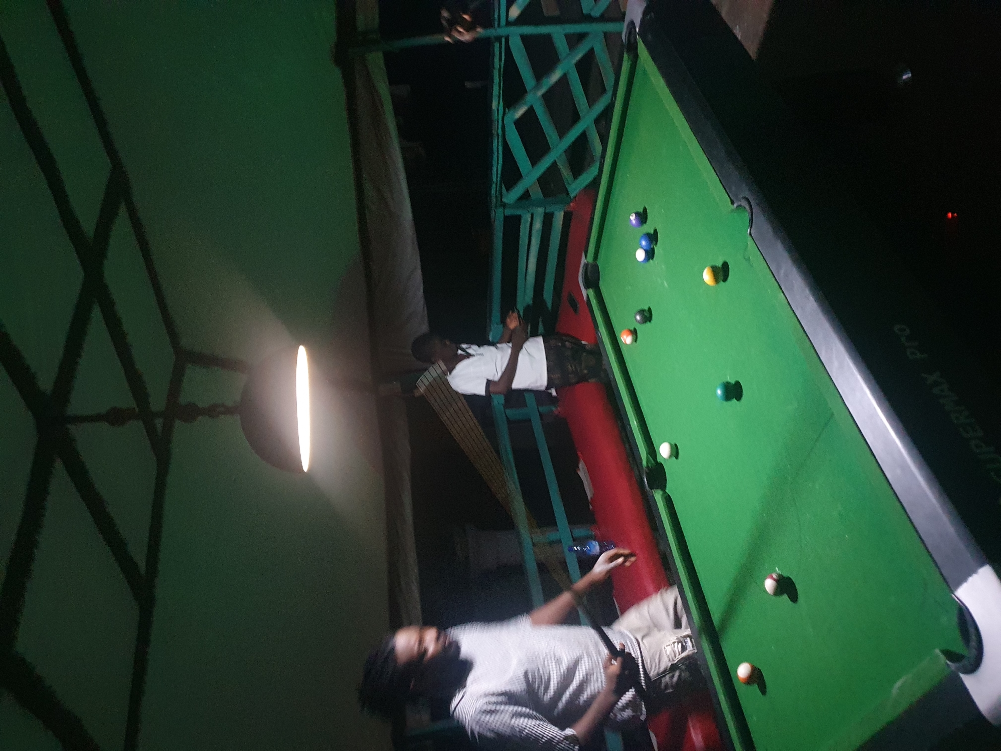 Two men are playing billiards on a green felt pool table at night, likely in Lokoja, Nigeria. The table is lit by a single overhead lamp. One man, wearing a striped shirt and tan shorts, is on the left, holding a cue stick and looking towards the table. The other man, in a white t-shirt and camouflage shorts, stands on the right, also facing the table. Several billiard balls are scattered on the table, including a white cue ball, a solid yellow ball, a solid blue ball, and a striped ball. The background shows a dark, possibly outdoor or semi-outdoor setting with a wooden railing and a dark sky. The environment appears casual, possibly a bar or recreational area.