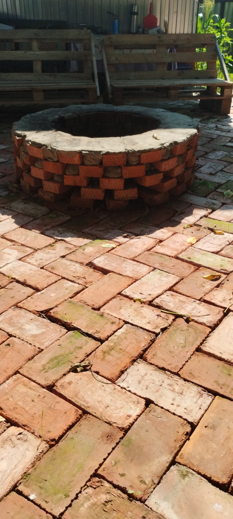 An outdoor scene in Yaroslavskaya, Russia, features a circular brick fire pit with a rough stone top, situated on a herringbone brick patio. Two rustic benches constructed from wooden pallets are positioned behind the fire pit. The environment appears to be a backyard or outdoor living space, with elements of greenery and sunlight casting shadows. There are no people visible in the image.