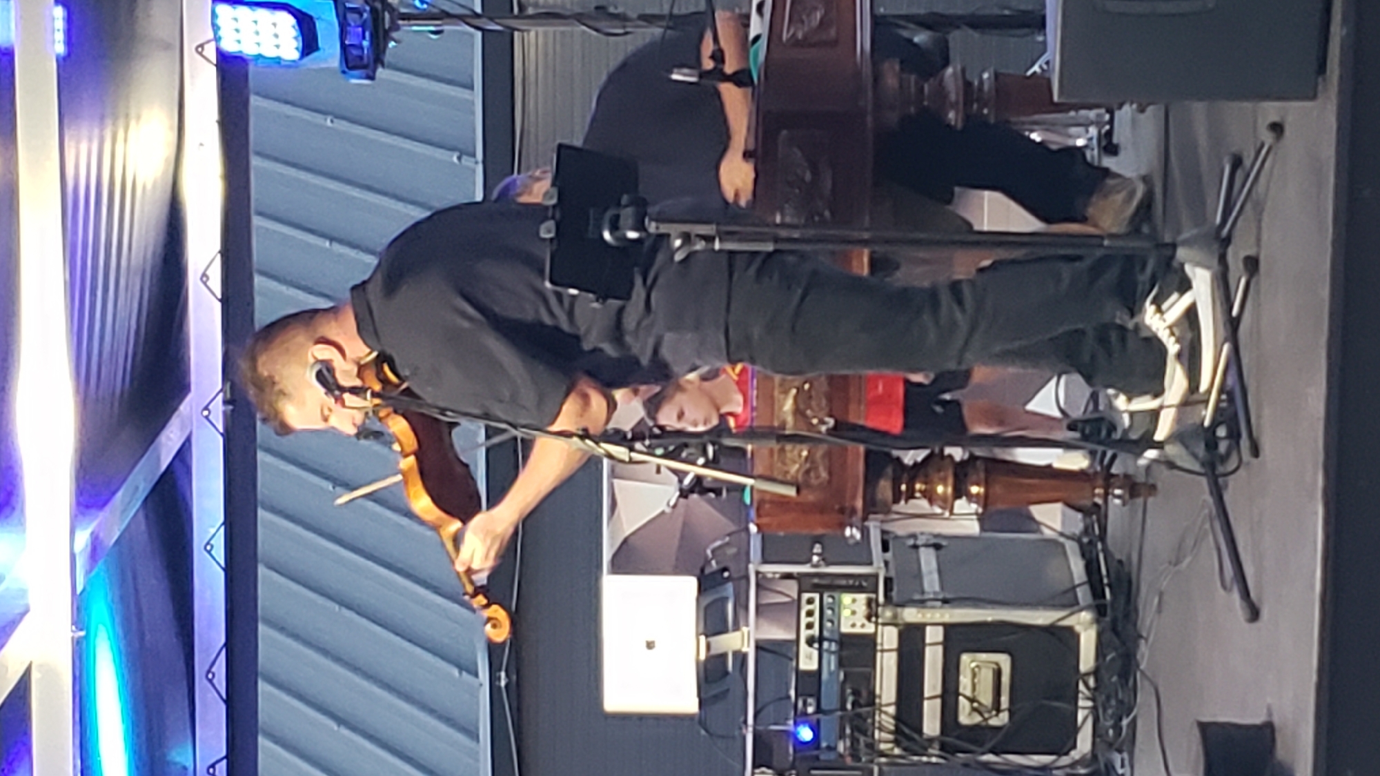 The image captures a live musical performance, likely outdoors or in a large venue, under a structure illuminated by blue lights. A man in a black shirt and pants is actively playing the violin, holding the instrument against his shoulder and drawing the bow across the strings. His eyes are focused on his playing, and a microphone is positioned near his mouth. To his left, another person is partially visible, also seated. In the background, a young boy in a red shirt is watching the performance, and a laptop sits on a stand. Various pieces of audio equipment, including speaker cabinets, rack-mounted electronics, and cables, are set up on stage, suggesting a professional musical setup. The overall environment appears to be a stage with a dark floor and a metal roof structure above. This scene is located in Prešov, Slovakia.