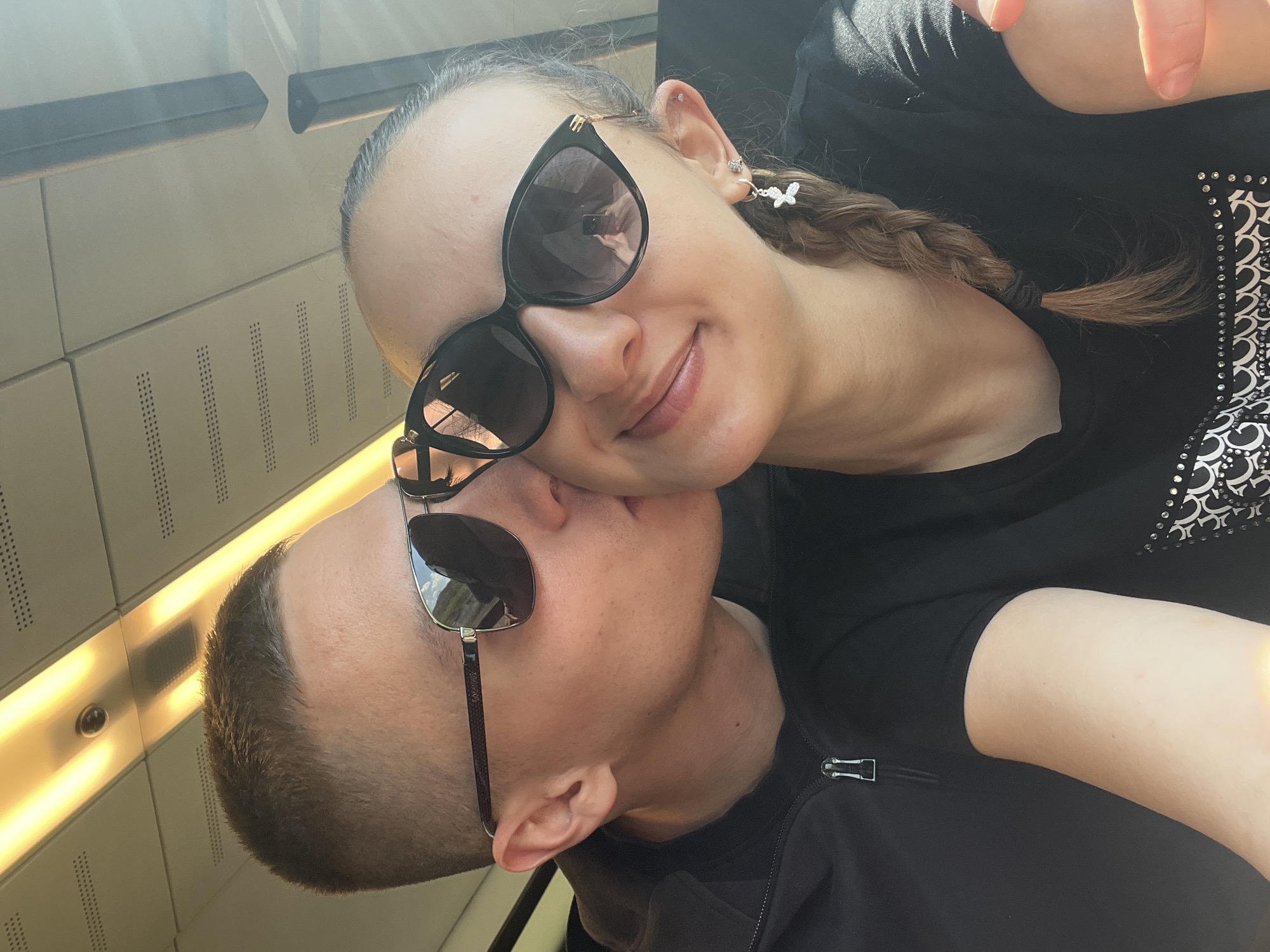 A selfie shows a woman and a man closely positioned, possibly in an affectionate pose. The woman has light skin, a braid, and wears butterfly earrings and dark sunglasses. The man has short hair and also wears dark sunglasses. Both are dressed in black clothing. The background suggests an interior space with white paneling and a lighting fixture. The location is Rehlingen, Germany.