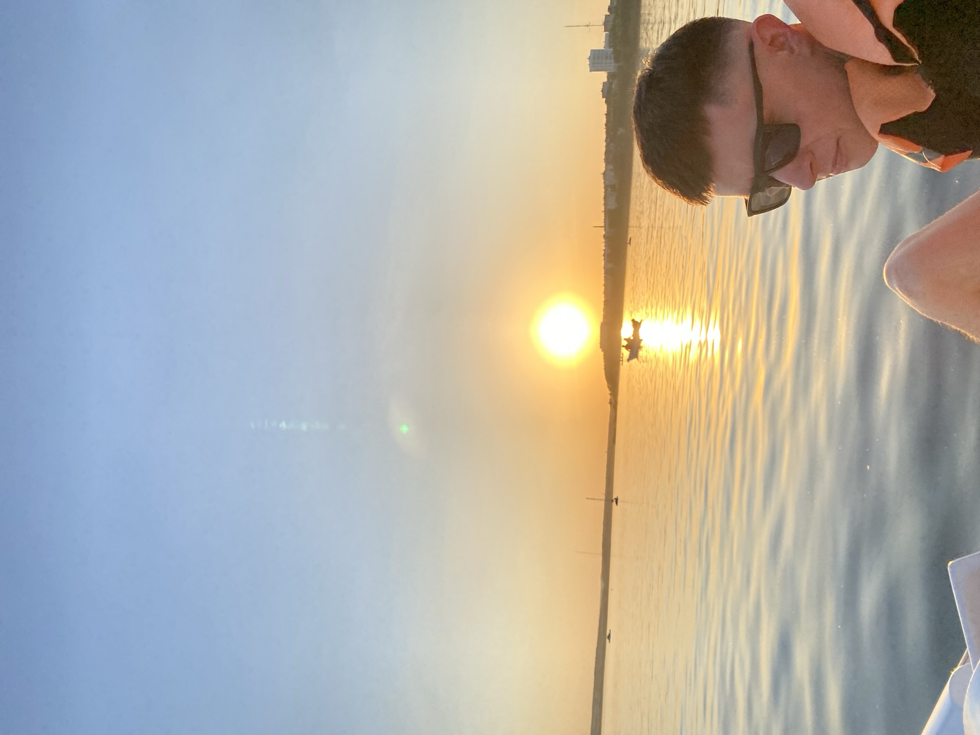 A man wearing sunglasses and a life vest is pictured on a boat at sunset, with the sun reflecting on the water, in Izhevsk, Russia. Buildings are visible in the distance. A few small boats or buoys are visible on the water. The sky transitions from blue to orange near the horizon.
