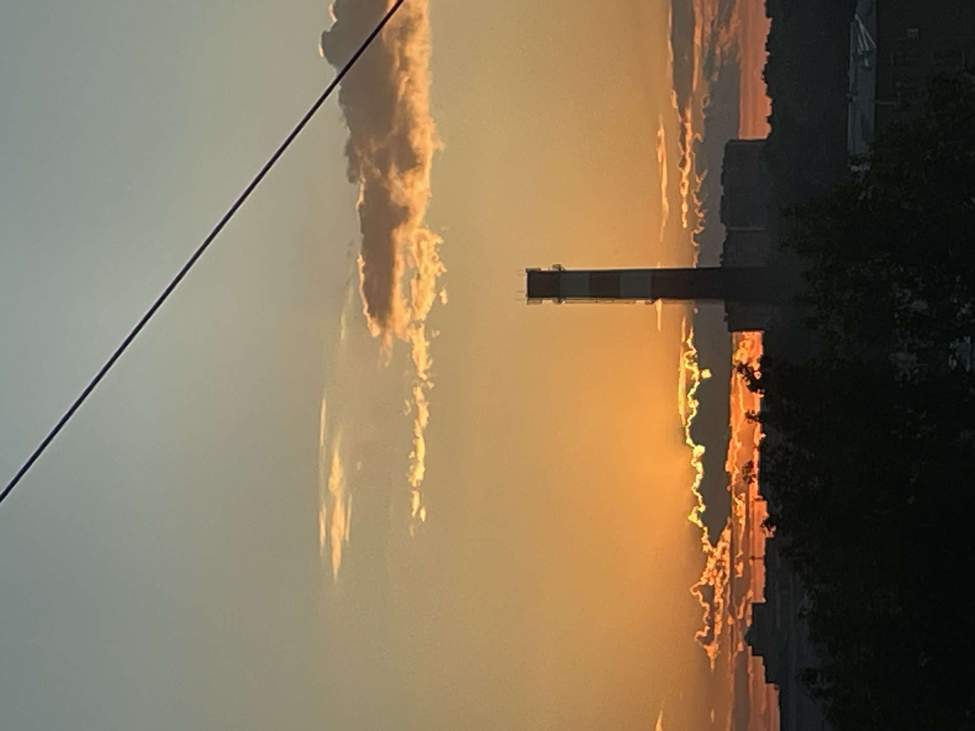 The image depicts a sunset scene over an urban or industrial landscape. The sky transitions from a light blue-grey at the top to a vibrant orange and deep yellow near the horizon, indicative of late afternoon or early evening. Clouds are scattered across the sky; those closer to the horizon are illuminated from below by the setting sun, displaying bright orange and yellow edges. The sun itself is partially obscured by clouds on the horizon, manifesting as a intense orange glow behind and between cloud formations.
A tall, slender industrial chimney or smokestack stands prominently in the middle ground, slightly to the right of the frame's center. It features distinct horizontal bands of alternating dark and light colors (appearing as a dark red/brown and white/light grey against the bright sky). Its top includes a cap-like structure.
Below the chimney and sun, a dark silhouette of a city skyline is visible on the horizon, composed of various buildings and structures. Further in the foreground, a dense, dark mass of trees and vegetation forms a prominent silhouette, obscuring the lower portions of the cityscape. In the bottom right corner, partially obscured by foliage, a building with a distinct roofline is silhouetted.
A thin, dark line, consistent with a power line or cable, traverses the upper portion of the frame diagonally from the upper left to the middle right. There are no individuals or discernible actions present in the scene. The location is an unspecified urban or industrial area in Unknown, Unknown.