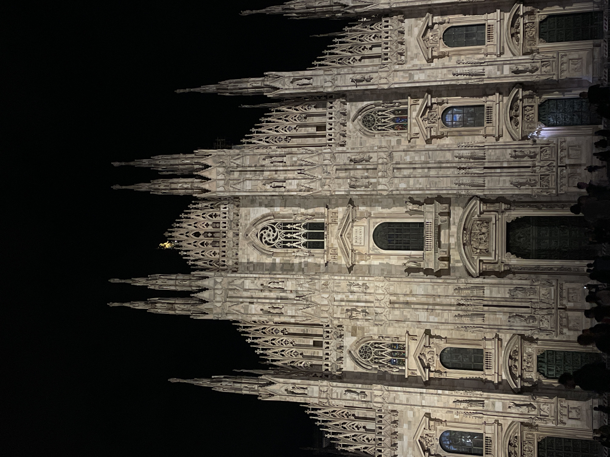 The image shows the Milan Cathedral, also known as the Duomo di Milano, at night. The facade is intricately detailed with spires, statues, and ornate window frames. The building's structure is made of light-colored stone. A group of people are visible on the right side of the frame. The sky is black, indicating the time is at night.