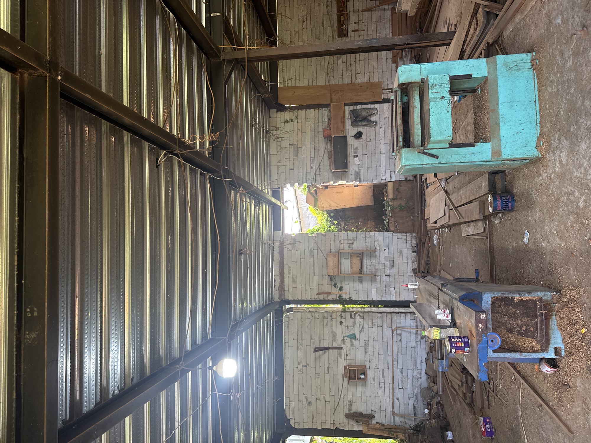 This image depicts the interior of a workshop, likely in Tampaksiring, Indonesia, characterized by its rustic and utilitarian nature. The ceiling is constructed from corrugated metal sheets supported by exposed steel beams, with electrical wiring and a bare lightbulb hanging from above. The walls are made of light-colored, horizontally stacked bricks.
In the foreground, to the left, is a large, blue woodworking machine with a flat, wooden workbench on top. Several cans and bottles of liquids, likely adhesives or finishes, are placed on the workbench. Wood shavings are scattered on and around the machine, indicating recent use. To the right, a bright turquoise industrial planer machine stands, also surrounded by wood shavings.
The floor is made of packed dirt, littered with wood scraps, planks, and various tools. In the background, an opening in the wall reveals an overgrown, outdoor area with lush green vegetation. The overall impression is one of an active but unrefined woodworking space, possibly a small business or individual artisan's workshop.