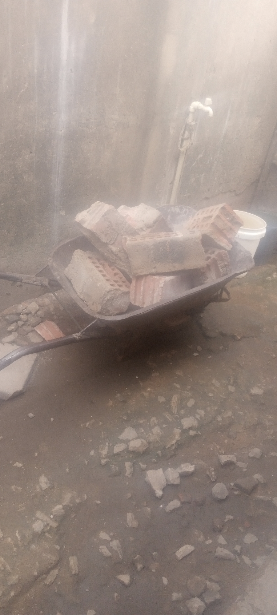 The image depicts a construction site in Sabon Tasha, Nigeria. A wheelbarrow is filled with red bricks, partially obscuring a white bucket in the background. A water tap is affixed to the wall. Debris and loose stones cover the ground. The scene appears to be outdoors.