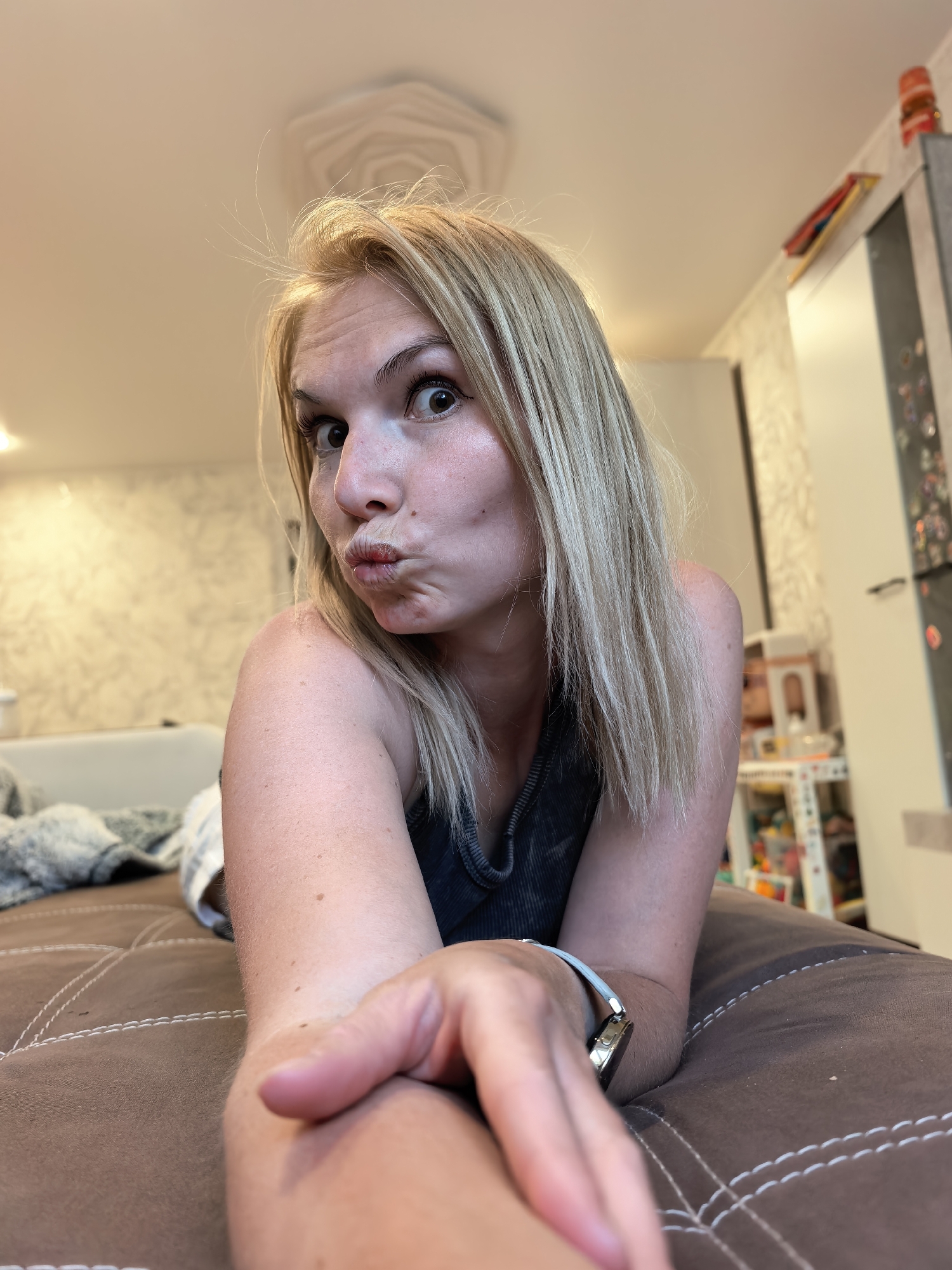 A woman with blonde, shoulder-length hair is lying on a brown, upholstered surface. She is wearing a dark tank top and a watch on her left wrist with a light-colored band. Her face is angled toward the camera, and she is making a pouting expression. The background shows a room with light-colored walls with a patterned design. A gray sofa is visible in the background, along with a white refrigerator with magnets and a shelf or storage unit containing children's toys. The location is in Sterlitamak, Russia.