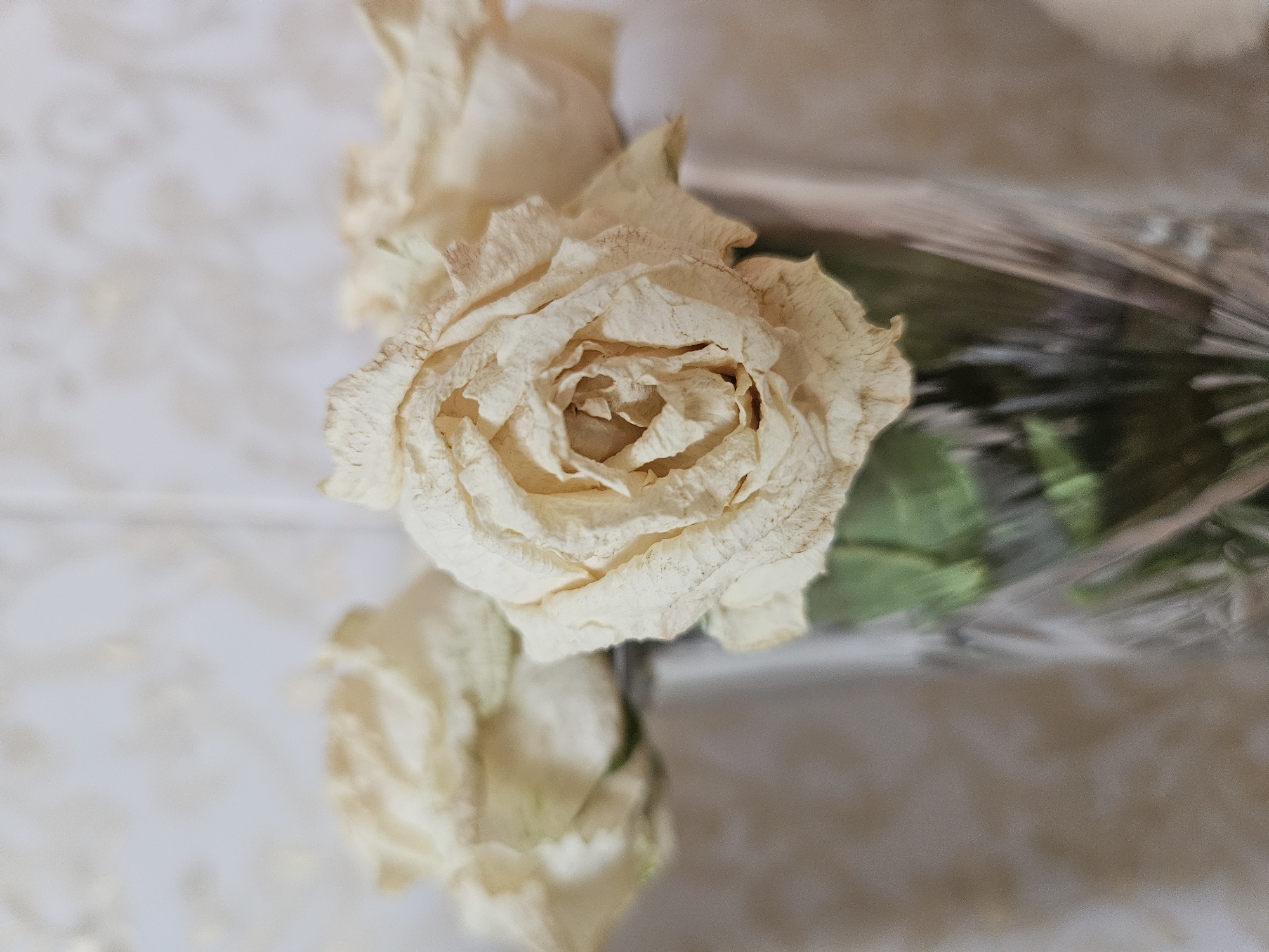 A close-up, shallow depth-of-field photograph shows a bouquet of three dried, pale yellow roses in a clear, crystal vase. The roses are wilting, with their petals dry and slightly curled, some showing brown edges. The vase is textured and appears to be made of cut glass, reflecting light. The background is a soft, blurry pattern of light beige and white, suggesting a wall or fabric. The scene is indoors, with no indication of the city of Andijan City, Uzbekistan.