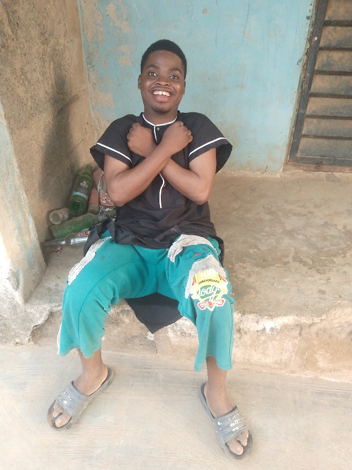 A smiling young man sits cross-legged on a concrete step, arms crossed over his chest in an "X" shape. He wears a black short-sleeved shirt with white trim and green cropped pants with patches. Gray sandals are on his feet. Behind him, against a weathered blue wall, sit several glass bottles. A slatted window is to the right, and the surrounding area appears to be an outdoor or semi-outdoor space.