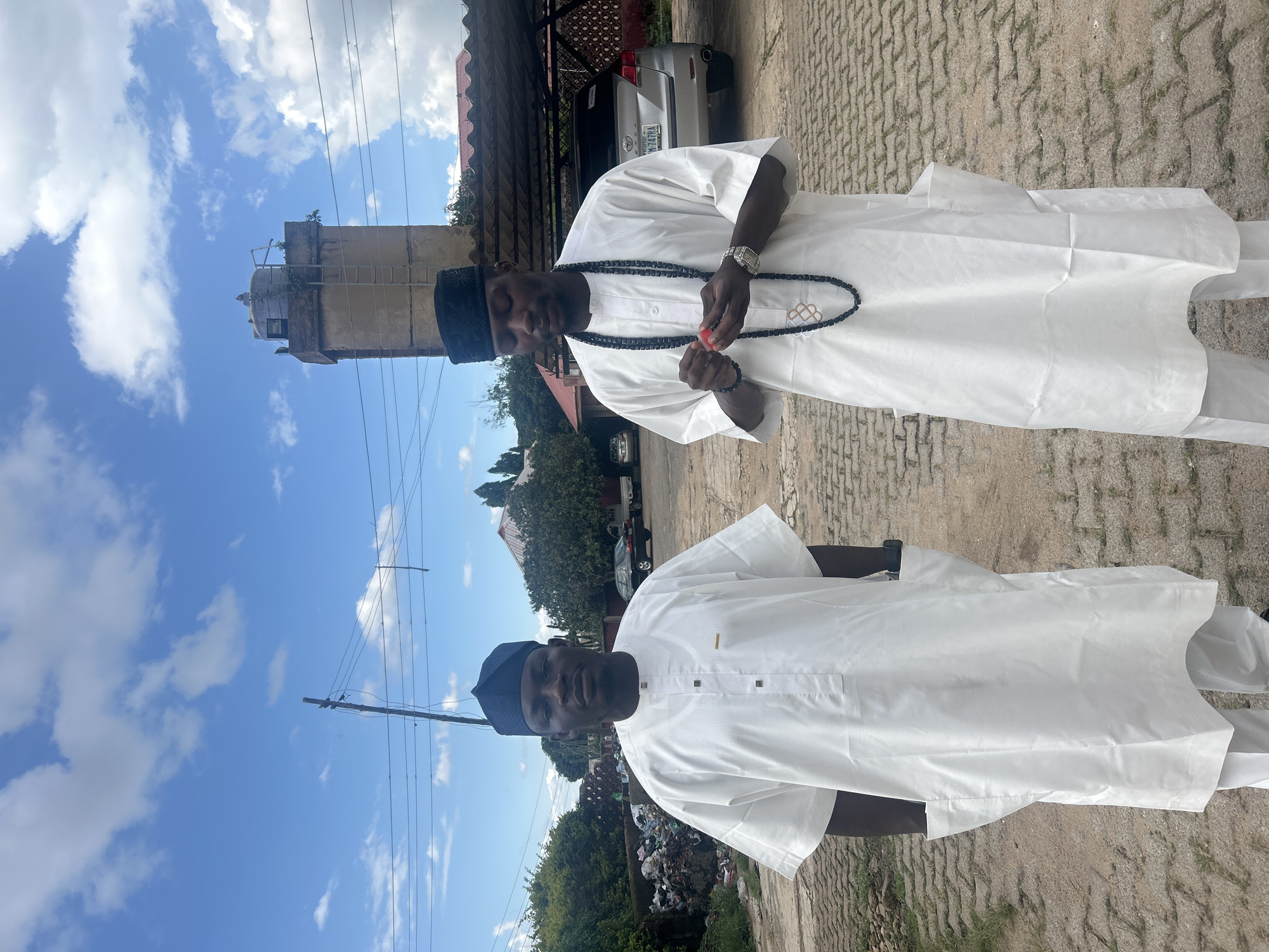 Two men stand outdoors in what appears to be a semi-urban or industrial area in Jos, Nigeria. The man on the left is facing the camera directly, wearing a white traditional outfit with a black cap. The man on the right is looking down at something red in his hands, also wearing a similar white outfit and a black cap, and a string of dark beads around his neck. In the background, there is a large concrete structure resembling a water tower, some parked cars, a pile of refuse, and trees under a cloudy sky. The ground is paved with interlocking bricks. The presence of the water tower and the general landscape suggest a location within Jos.