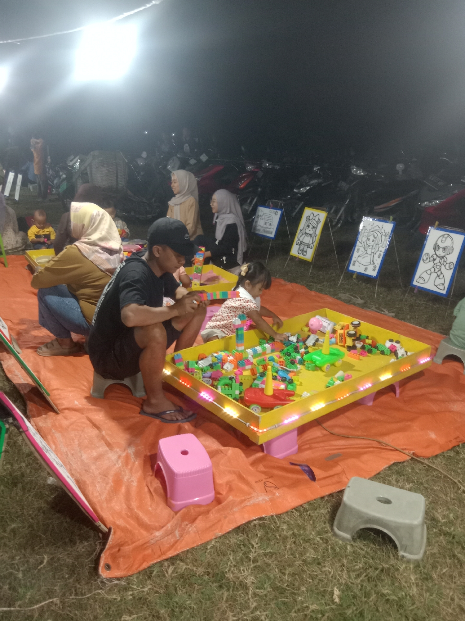 In Stiaji, Indonesia, at an outdoor night market, a family gathers around a bright yellow toy table filled with colorful building blocks and toys. A young girl in a patterned dress is actively engaged, reaching into the toy bin. Beside her, a man wearing a black t-shirt and shorts squats, intently examining a toy. Further back, a woman in a brown top and hijab is also seated, watching the activities. Other individuals, including a baby in a yellow shirt, are visible in the background, suggesting a communal gathering. The scene is illuminated by bright overhead lights, casting a stark glow on the orange tarp laid out on the grass. In the background, numerous motorcycles are parked, indicating a popular event. Educational posters featuring cartoon characters are displayed, adding to the festive atmosphere.
