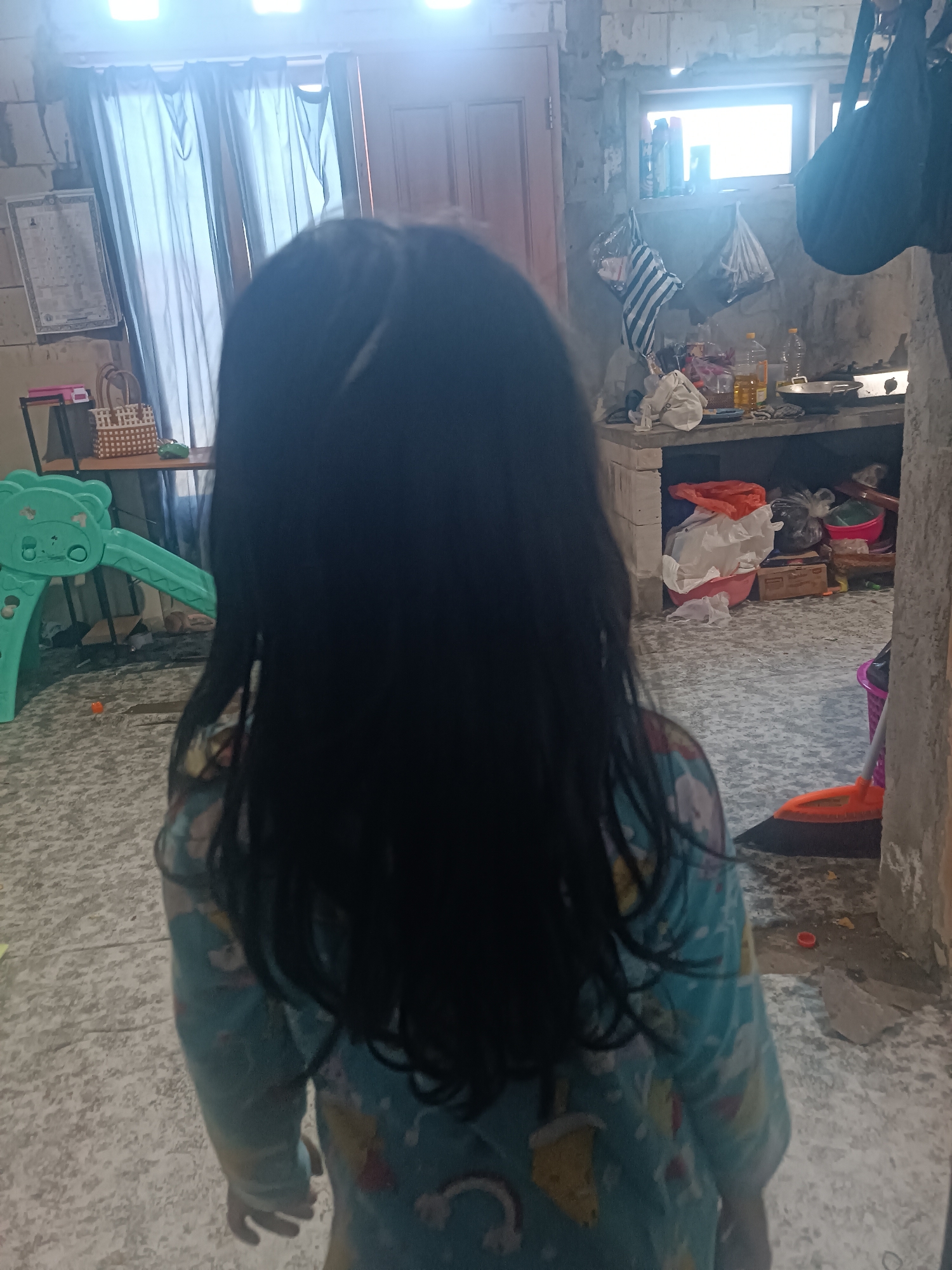 In Tabanan, Indonesia, an image taken from behind shows a young child with long, dark hair wearing a blue pajama top with a cartoon print. The child is standing on a textured floor in what appears to be an unfinished interior space. To the left, a green plastic slide is visible, suggesting a play area. The background includes a wall with exposed brickwork, a window with a bright light emanating from it, and various household items and clutter, including bags, bottles, and cleaning tools like a broom. The overall environment suggests a utilitarian or construction zone within a home.