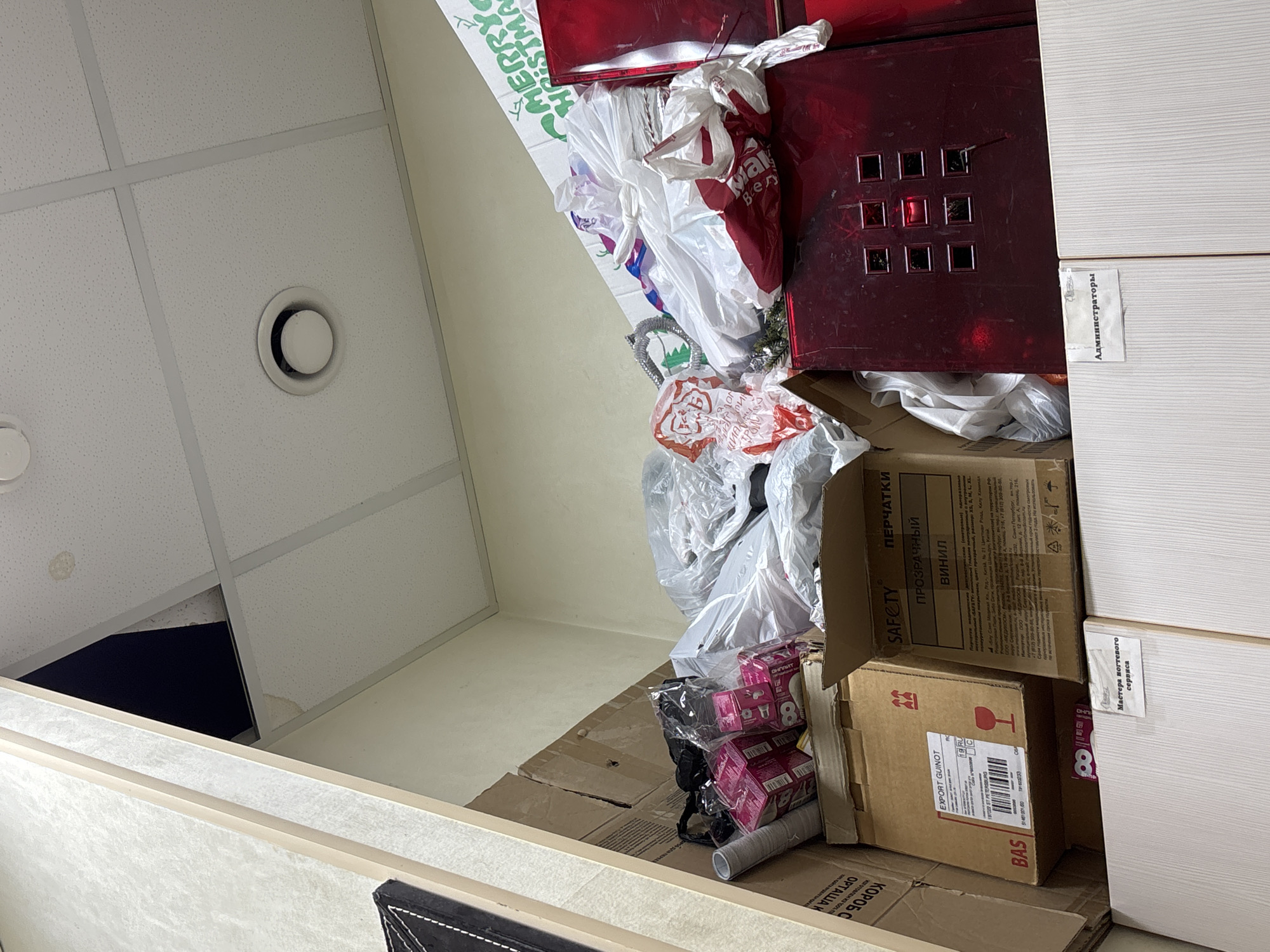 In the city of Ladozhskoe Ozero, Russia, a cluttered storage space is depicted. The image focuses on a dense collection of various items piled high, obscuring much of the room. Dominating the scene are numerous cardboard boxes, some open and others sealed, containing what appears to be merchandise or supplies. Among these boxes are several plastic bags, some filled and tied, others appearing to be empty.
Prominently displayed is a large red cabinet or container, its surface reflective and featuring a grid of nine square, dark openings. Various labels are visible on the boxes, including one that reads "EXPORT GUINOT," and another in Russian that translates to "PERCHATKI" (Gloves) and "VINIL" (Vinyl). Another label indicates "Mastera podtegovogo servisa" (Master of the flooring service) and a sign that reads "Администраторы" (Administrators).
A portion of the ceiling is visible, composed of white acoustic tiles with a ventilation grille. A dark opening is present in the ceiling to the left, suggesting a possible access point or structural anomaly. The overall impression is one of disarray and a significant accumulation of stored goods in a commercial or administrative setting.