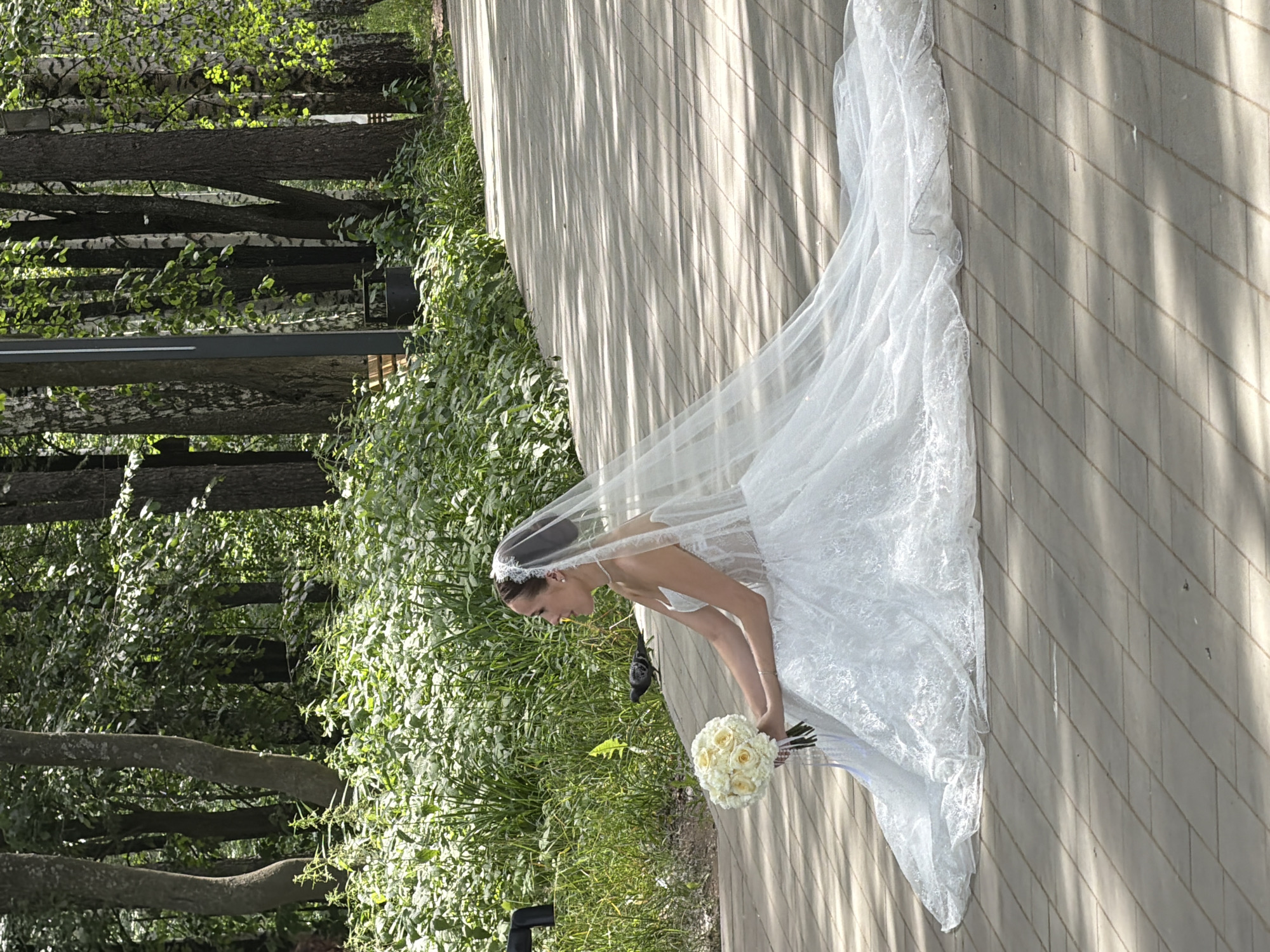 A bride in a white wedding dress and veil sits on a paved path in a park, holding a bouquet of yellow roses. She is looking to her right, and a pigeon is visible on the path in front of her. The park has tall trees and lush green foliage. The paved path is bathed in dappled sunlight, creating a pattern of light and shadow. The overall scene suggests a peaceful outdoor setting for a wedding photoshoot.