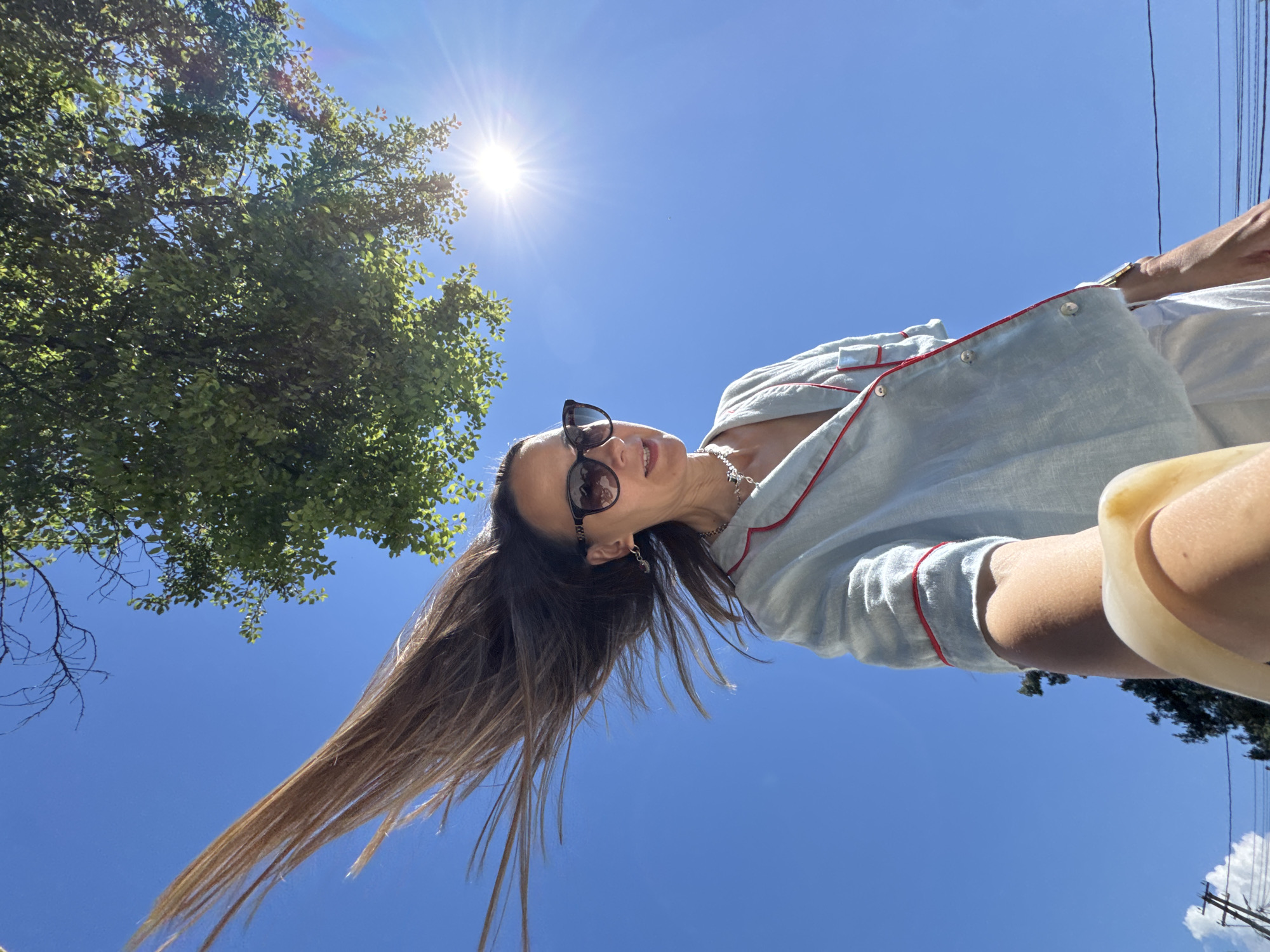 A low-angle, wide-shot, full-body selfie captures a woman with long, brown hair, partially obscured by her own arm extending outward, as she stands outdoors under a bright, sunny blue sky. Her hair is being swept dramatically to the left by the wind. She is wearing sunglasses, a light blue pajama-style top with red trim, and white bottoms. A necklace with a pendant is visible around her neck. The sun, a bright, lens-flared star, is positioned in the upper right quadrant of the image, casting a warm glow. Green foliage from a tree branches down from the top of the frame, also bathed in sunlight. In the background, distant power lines and the tops of trees are visible against the clear sky. The scene appears to be set in an urban or suburban environment, possibly a street in Sochi, Russia, given the presence of power lines and leafy trees. The overall impression is one of a casual, spontaneous moment captured on a clear, windy day.