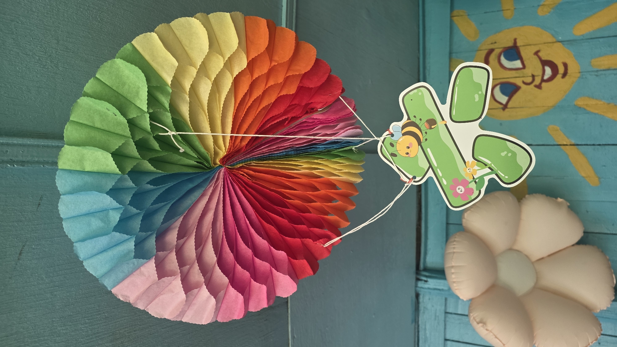 An overhead shot captures a vibrant, rainbow-colored paper fan decoration suspended from a teal-colored ceiling. Below the fan, a green cutout of a frog with a cartoon bee perched on its back hangs by white strings. To the left of the frog cutout is a soft, light pink flower-shaped pillow. In the background, a portion of a cheerful yellow sun mural with a smiling face is visible on a teal-colored wooden wall. The overall scene suggests a child-friendly environment, possibly a playroom or a classroom, in Tokmok City, Kyrgyzstan.
