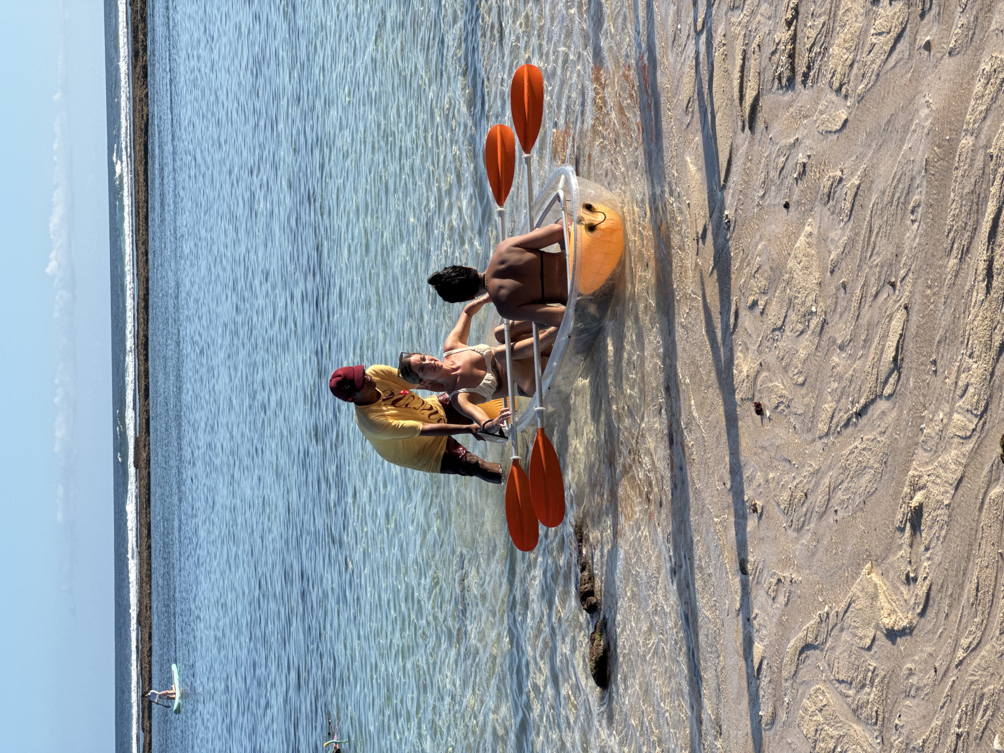 The image captures a sunny coastal scene in Kuta Selatan, Indonesia. A clear kayak, containing two people, is positioned partially on the sandy beach and partially in the shallow, clear water. The beach has a textured surface of wet sand.
The kayak contains two women. One is reclining and appears to be wearing a light-colored bikini. The other woman is seated behind her, wearing a dark swimsuit. Beside the kayak, a man in a yellow t-shirt, dark shorts, and a maroon cap is assisting or interacting with the occupants.
Four orange paddles are placed around the kayak. In the background, the water stretches to the horizon, with a thin line of rocks. A person on a paddleboard can be seen far in the distance. The sky is a pale blue.