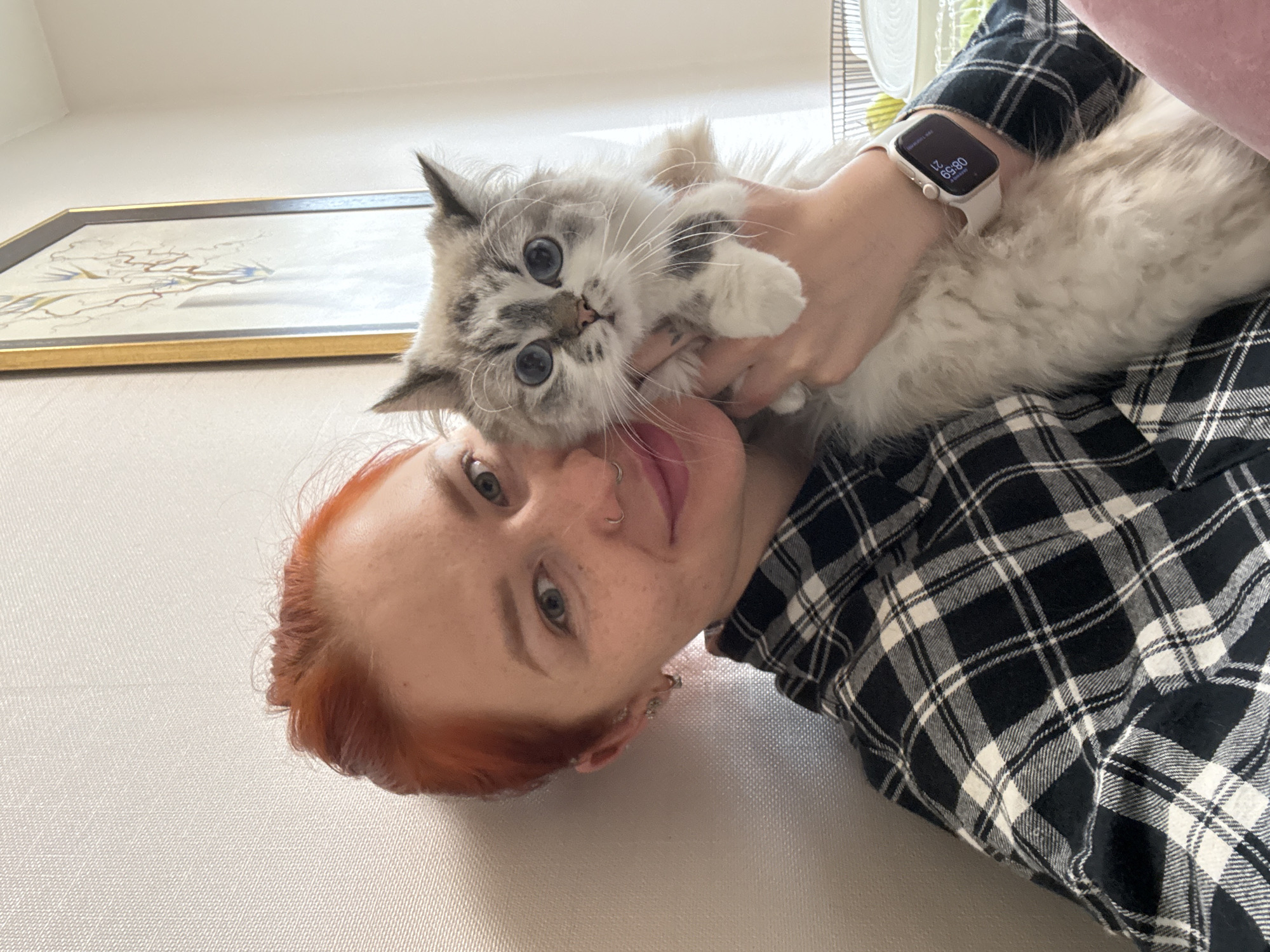 An intimate, selfie-style portrait captures a person with red hair cuddling a fluffy cat indoors. The scene is set in what appears to be a home in Rostov-on-Don, Russia.
The main subjects are a young person, likely female, with vibrant red hair, freckles, and light blue eyes, and a domestic cat. The person is seen from the chest up, lying down and looking towards the camera with a gentle smile and relaxed expression. They are wearing a black and white plaid shirt. Their left arm is wrapped around the cat, holding it close. On their left wrist, a white Apple Watch is visible, displaying the time "08:59" and a notification for "Birthday List."
The cat, held securely, also looks directly at the camera with large, striking blue eyes, appearing wide-eyed and perhaps a little surprised or curious. It has soft, light grey and white fur with darker tabby markings on its face and paws, and prominent whiskers.
In the background, a light-colored wall is visible, with a framed picture or painting featuring an abstract, delicate design in muted gold and blue tones. Part of a window or a white-bladed fan can be seen in the upper right. The lighting suggests daytime. The overall impression is one of affection and companionship between the owner and their pet.
