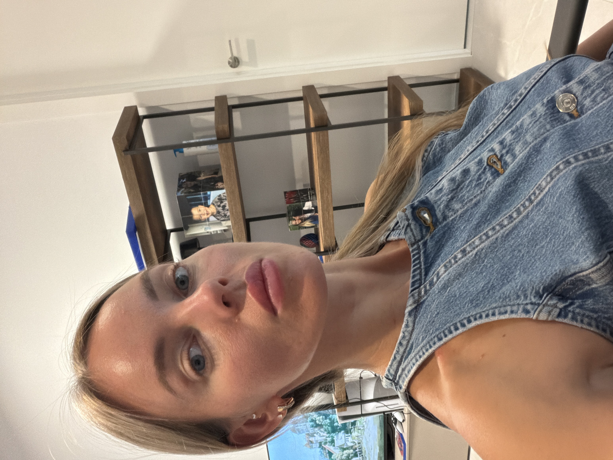 A woman with blonde hair and blue eyes, wearing a denim outfit, is taking a selfie in a well-lit room. She is positioned in the foreground, with a shelf unit containing framed photos and bottles behind her. A television screen displaying a landscape image is visible to her left. The environment appears to be a modern interior, possibly a home or office, with a neutral color palette. The scene is captured from a close-up perspective, focusing on the woman's face and upper body.