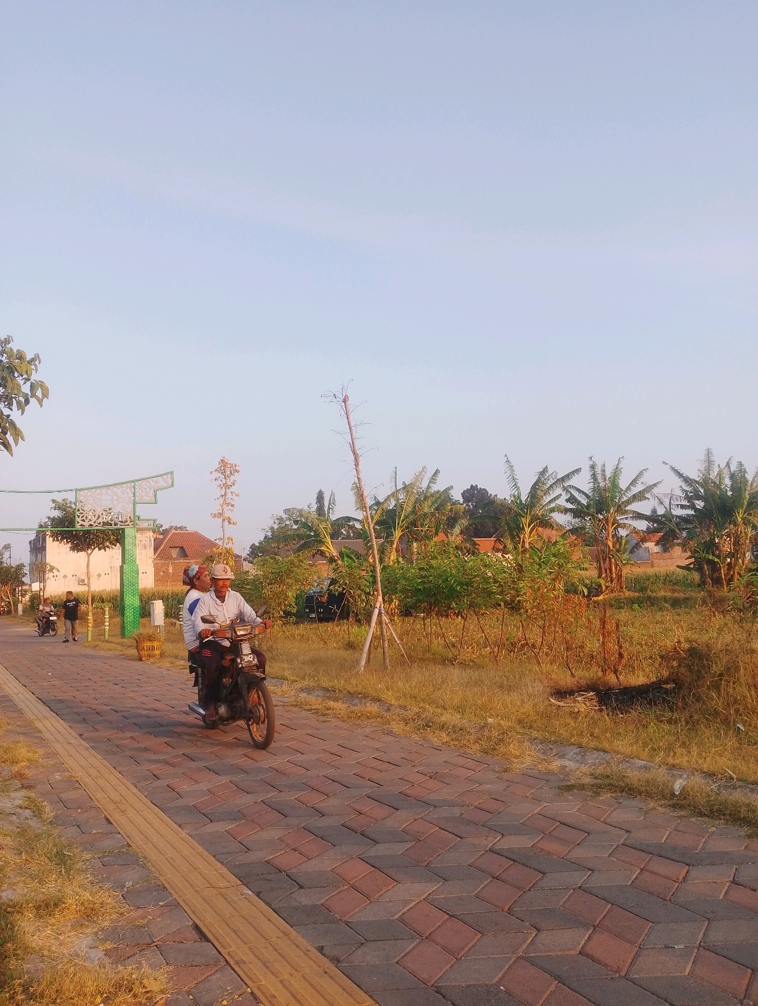 The image depicts a scene on a paved walkway in Probolinggo, Indonesia. In the foreground, a motorcycle carries two individuals, a man and a woman, riding along the brick-patterned path. The man is driving, and the woman is seated behind him. The walkway has a tactile paving strip running alongside it. In the background, there is an ornate green archway, houses with tiled roofs, and lush vegetation including banana trees. The sky is clear and light blue, suggesting it is daytime. Several other people are visible in the distance, walking or riding motorcycles on the same walkway. The overall atmosphere is tranquil and suggests a residential or recreational area.