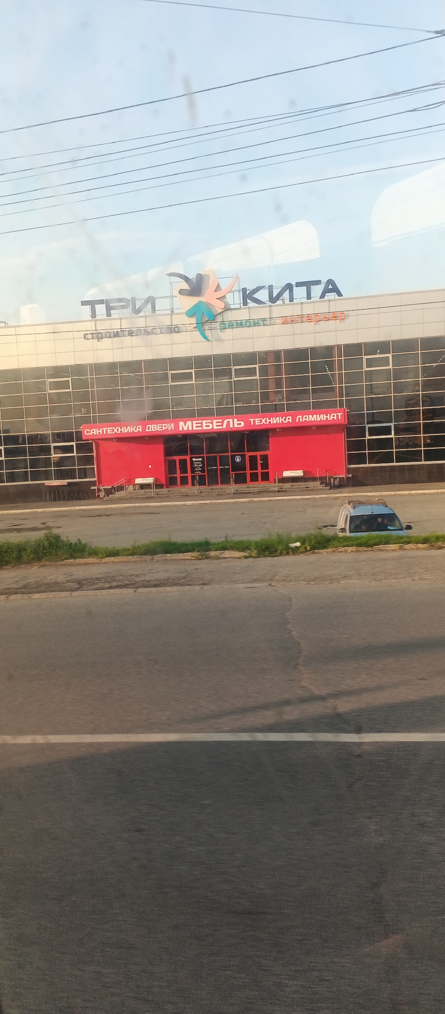 The image shows a building with a large sign that reads "ТРИ КИТА" (Three Whales) in Cyrillic. Below this, smaller text says "строительство ремонт интерьер" (construction, repair, interior). A red section of the building displays another sign with more Cyrillic text: "САНТЕХНИКА ДВЕРИ МЕБЕЛЬ ТЕХНИКА ЛАМИНАТ" (plumbing, doors, furniture, appliances, laminate). The building has a glass facade and a red entrance. To the right, a silver minivan is parked on the grass. The scene is likely a commercial area in Izhevsk, Russia, possibly a home improvement or furniture store. The image is taken from a slightly elevated perspective, possibly from a vehicle.