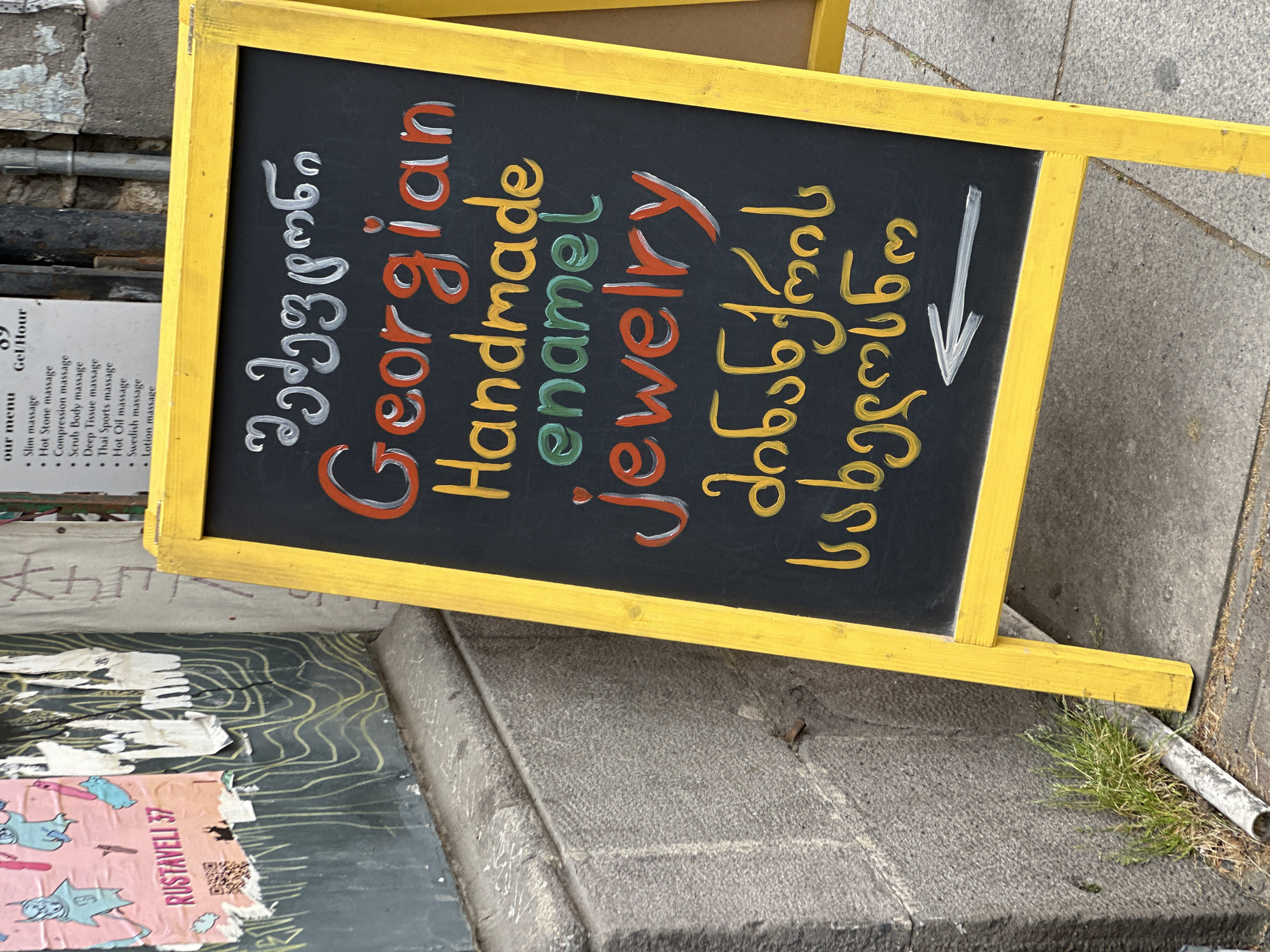 The image shows a close-up of a yellow-framed sign on a sidewalk, likely in Tbilisi, Georgia, indicated by the Georgian text on the sign and the "Rustaveli 37" address sticker visible below it. The sign advertises "Georgian Handmade Enamel Jewelry" and has a downward-pointing arrow, presumably directing customers to a shop. To the left of the sign, a partially visible menu lists various massage services. The sidewalk is concrete, with grass growing near the edge. A poster is visible at the bottom of the frame with a QR code and the text “RUSTAVELI 37”.