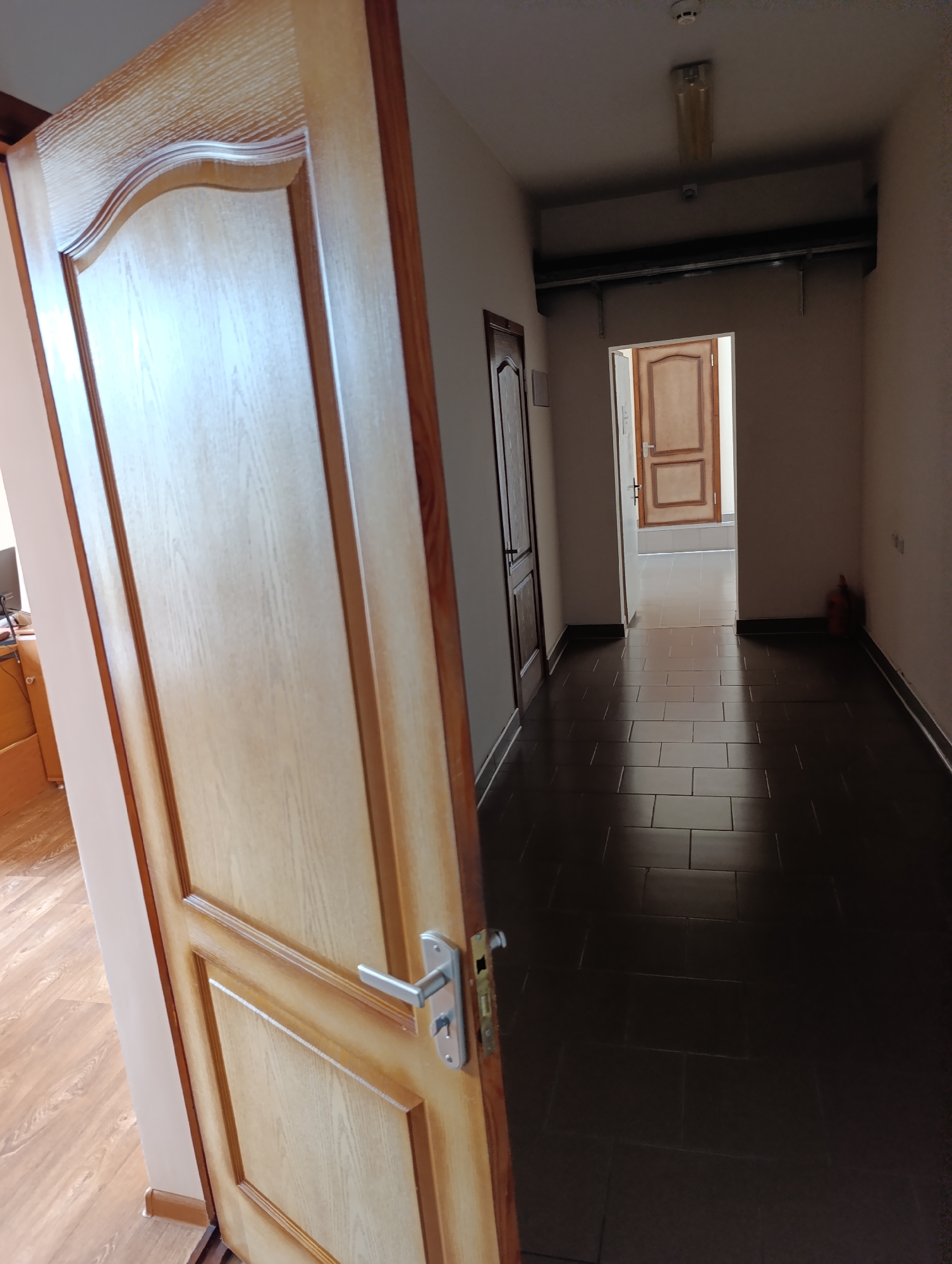 The image shows an interior hallway with dark tiled flooring. A light-colored wooden door with a silver handle is open on the left side of the frame, dominating the foreground. In the background, another doorway leads to a brightly lit room with a light-colored door. A red fire extinguisher is visible against the wall on the right side of the hallway. The lighting suggests an indoor environment. The scene is likely located within the Dubăsari town council building in Dubăsari, Moldova.