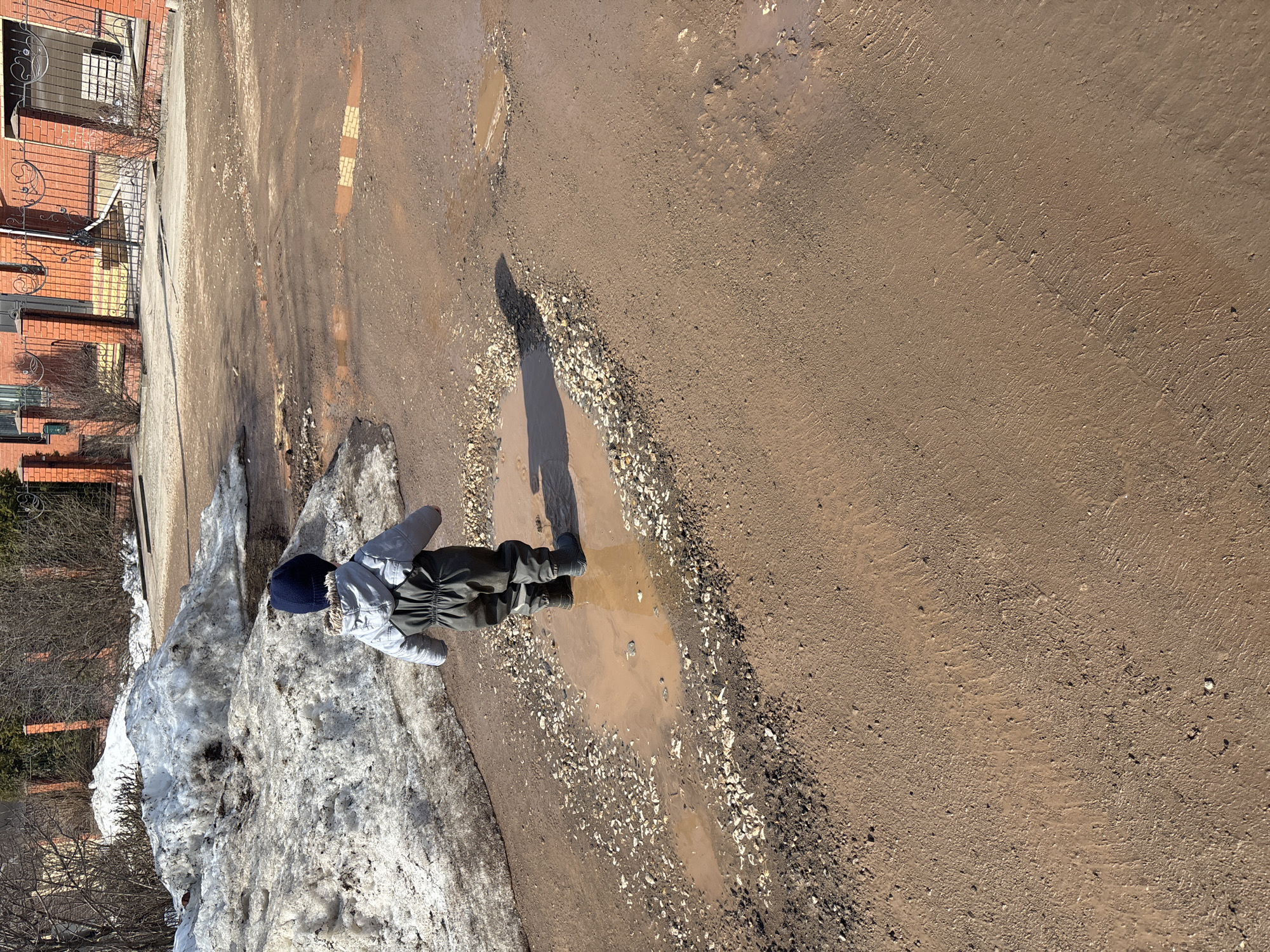 A child stands in a muddy puddle along a dirt road in Ферма, Russia. To the left, there is a large snow pile near a fence, trees, and a brick building with ornate wrought iron detailing. The ground is muddy and rutted. The child is wearing a dark blue hat, a light-colored coat with fur trim, and dark pants with black boots. The child's shadow stretches across the puddle. The road surface is a mix of mud, gravel, and what appears to be compressed dirt.