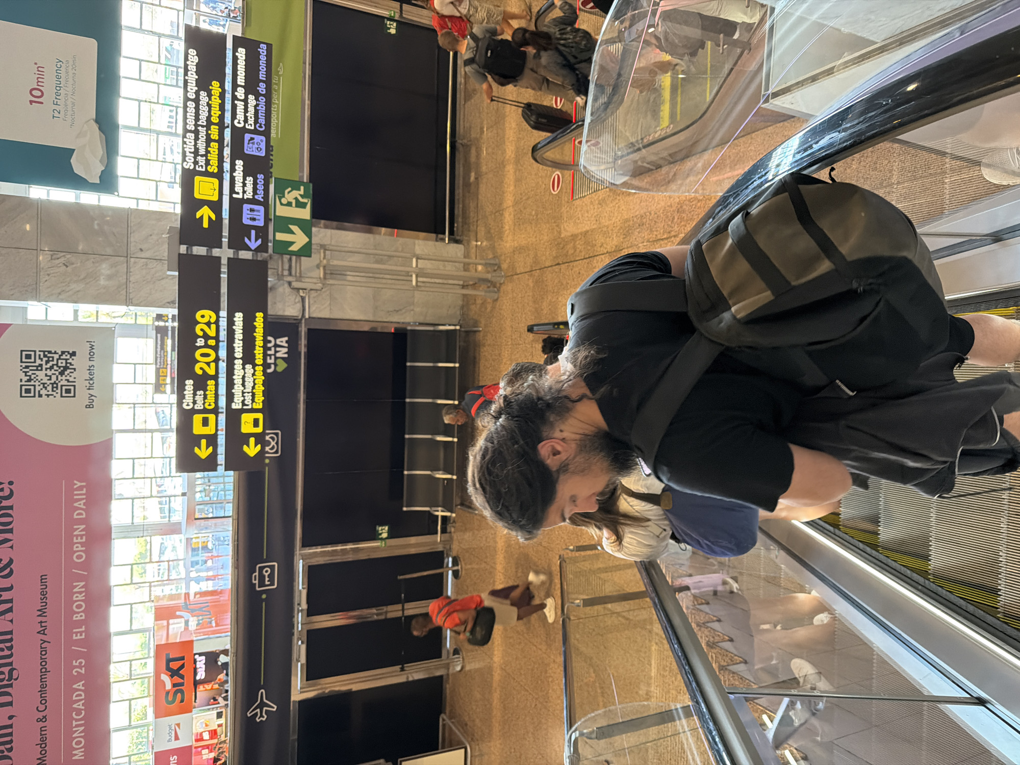 The image was taken at Barcelona-El Prat Airport in El Prat de Llobregat, Spain. It shows an indoor scene with multiple escalators and people navigating them. In the foreground, a person with long, dark hair and a beard is riding an escalator downwards, wearing a black t-shirt and a backpack. Other individuals are visible on escalators and walking in the background, some with luggage. Several directional signs are displayed, indicating information such as "Cintes" (belts), "Equipatges extraviats" (lost luggage), "Sortida sense equipatge" (exit without baggage), and "Lavabos" (restrooms). The overall environment appears to be a busy airport terminal.