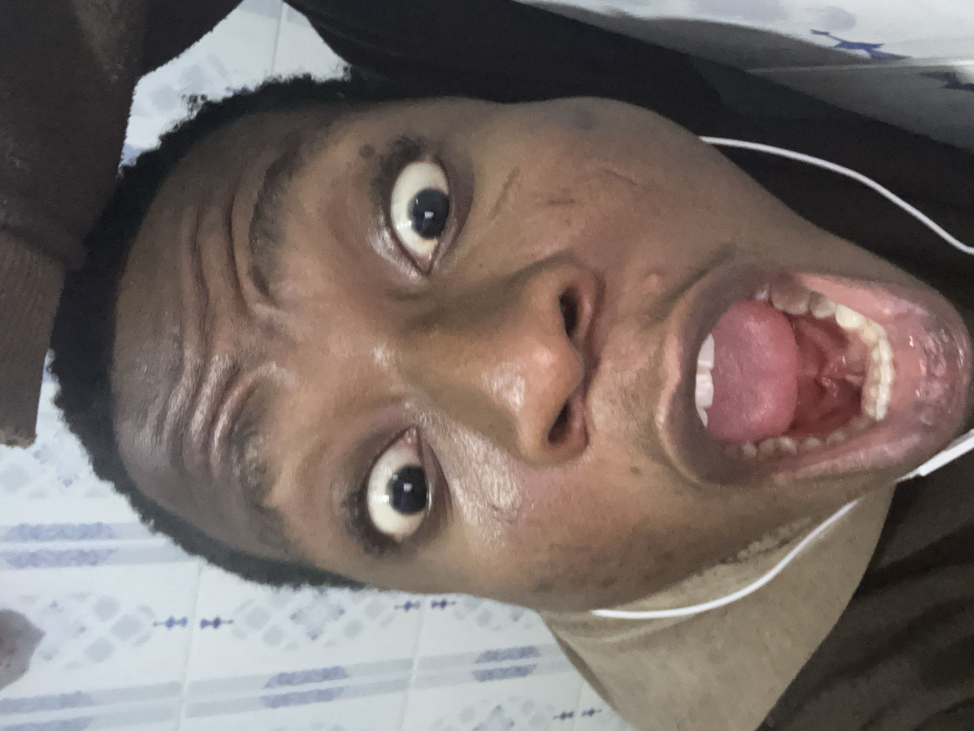 A close-up, angled view of a dark-skinned person's face with a wide-open mouth and eyes looking directly at the viewer. The person's tongue is visible and appears slightly raised. White earphones are visible alongside the person's neck. The background shows a tiled surface, presumably a wall, with a blue and white pattern. The location and city are unknown.