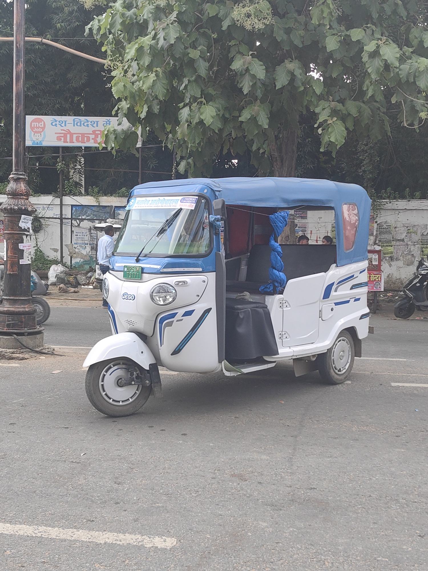 This image captures a white and blue auto-rickshaw parked on a paved road in Gorakhpur, India, under the shade of large green trees. The auto-rickshaw, with a "Wego" logo visible on its front, has its passenger door open. A person in a light-colored shirt and dark pants stands near the auto-rickshaw. In the background, a sign with Hindi text, including "md" and "देश-विदेश में नौकरी" (Jobs in India and Abroad), is partially visible, attached to a lamppost. There are several plastic bags and other debris on the side of the road. A black scooter is parked further back. The environment appears to be a street in a city with moderate vegetation.