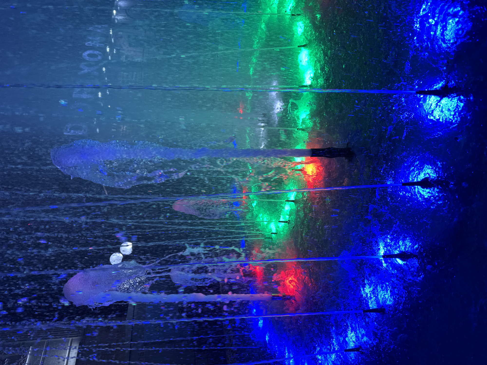The image depicts a colorful water fountain display at night. Water jets are illuminated by vibrant blue, green, and red lights, creating a dynamic visual effect. In the background, a building facade with illuminated signage, partially obscured by the water and mist, is visible. The signage appears to display the words "DIXON A". The scene is characterized by the interplay of light and water, with droplets and streams creating a sense of movement and energy. The overall impression is one of a modern urban landscape, likely a public space or entertainment venue in Tashkent, Uzbekistan, designed for aesthetic appeal and public enjoyment.