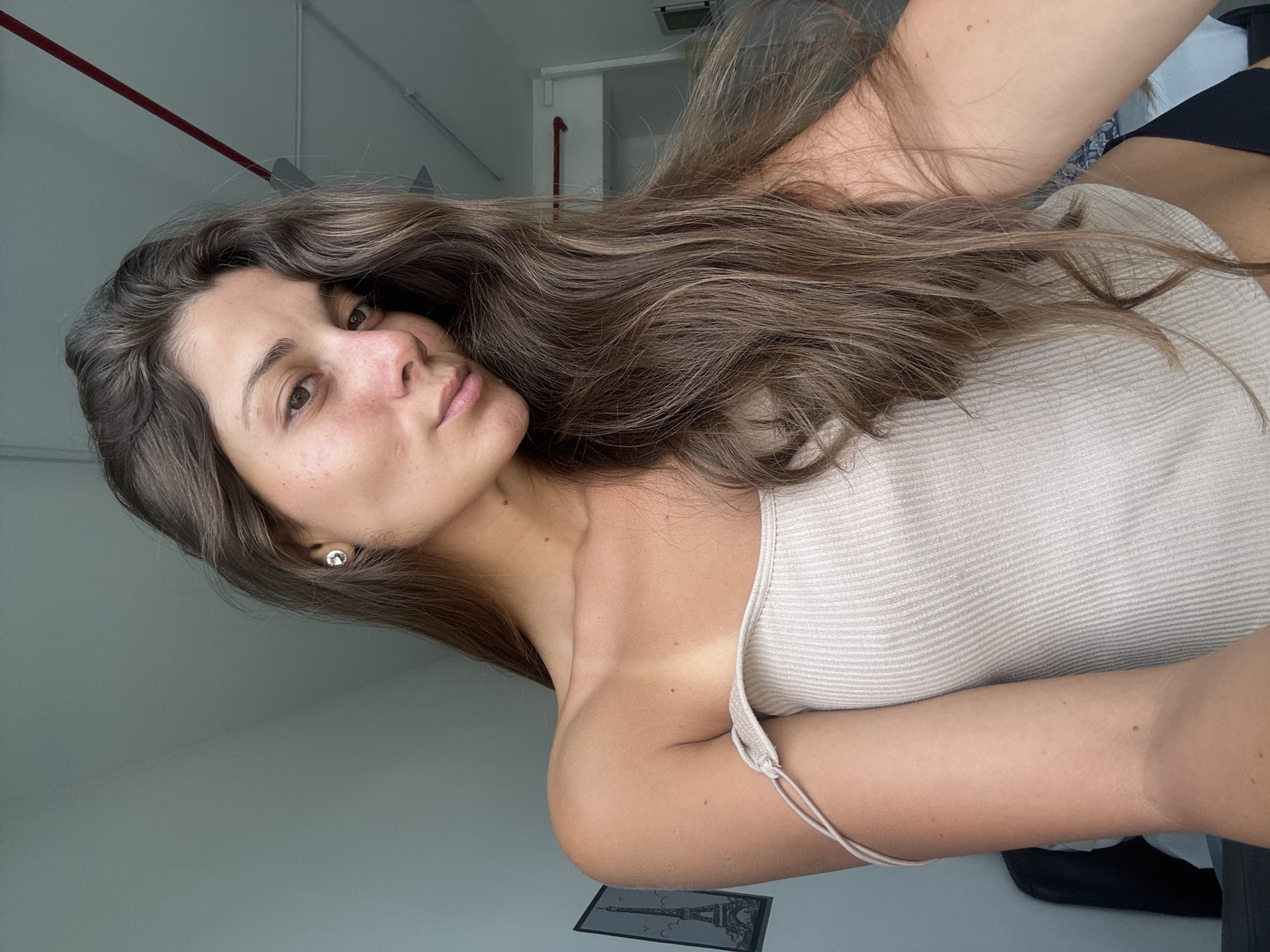 The image shows a young woman taking a selfie indoors, with the camera angled slightly downward and tilted. She has long, wavy brown hair that falls over her shoulders. Her skin tone is medium, with visible freckles on her nose and cheeks. She has brown eyes and is looking directly at the camera with a neutral expression, her lips slightly parted in a subtle pout.
She is wearing a light beige or off-white ribbed tank top with thin straps. A silver stud earring is visible in her left ear. The background consists of plain light-colored walls and a ceiling with a red pipe or conduit running across it. On a surface beneath her right arm, a small framed picture of the Eiffel Tower is visible. The lighting is bright and appears to be natural, indicating it is daytime. There is no discernible text in the image. The location context provided is Kuala Lumpur, Malaysia.