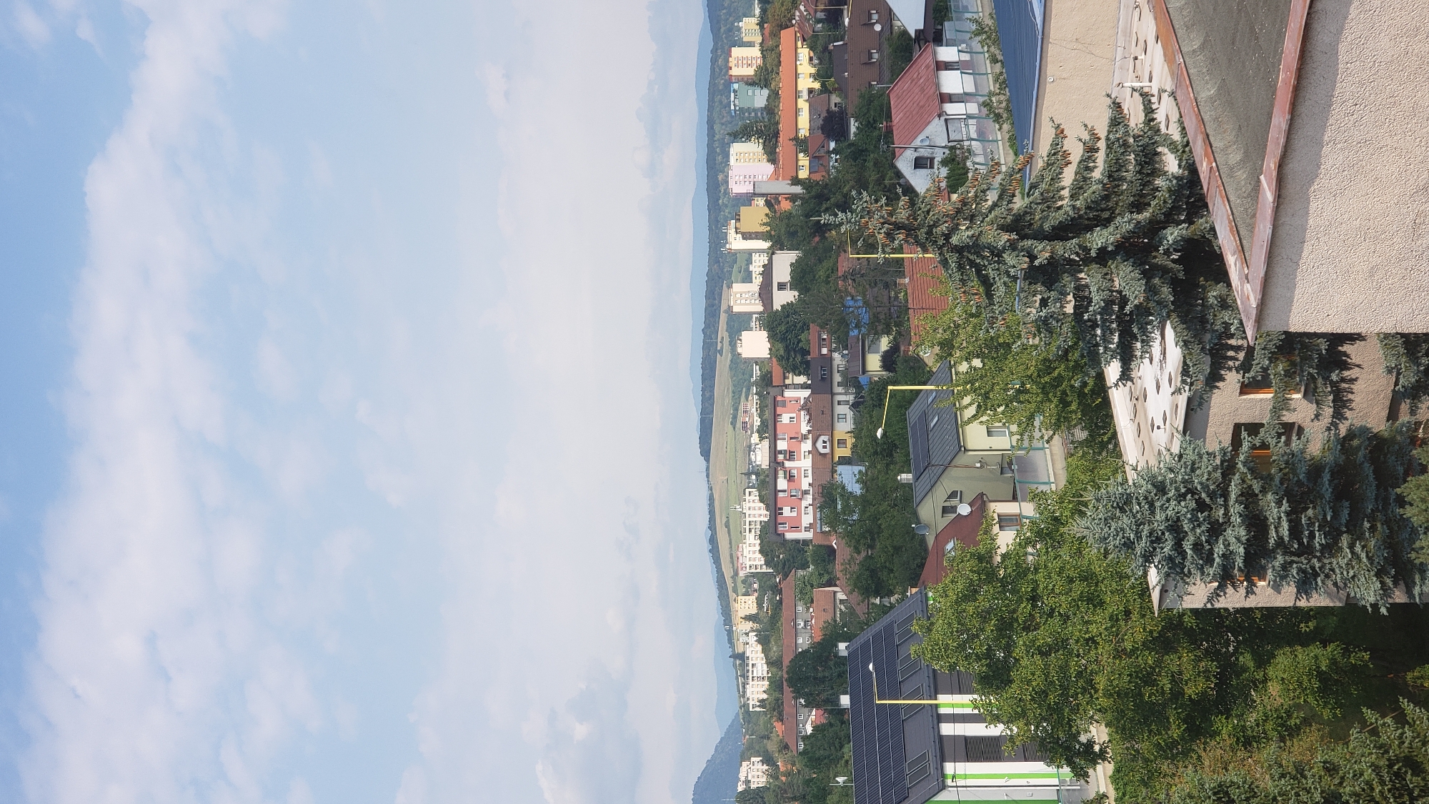 The image captures an elevated view of a residential area in Prešov, Slovakia, likely taken from a building. The foreground is dominated by the rooftops of several buildings, including one with a green and white facade and solar panels, and another with a textured, light-colored wall and a copper-colored trim on its roof. A tall evergreen tree with cones stands prominently in the middle ground, partially obscuring a light-colored building with two windows.
Beyond the immediate foreground, the scene opens up to a wider panorama of the city. A dense collection of houses and apartment buildings with various colored facades, including pink, yellow, and white, fills the mid-ground. Some of these buildings appear to be multi-story apartment blocks. Trees are interspersed throughout the cityscape, creating a natural contrast with the urban structures.
In the background, rolling hills and distant mountains form the horizon. The sky above is a pale blue with scattered white clouds, suggesting a clear or partly cloudy day. The overall impression is one of a typical urban or suburban landscape under daylight. There are no people visible in the image, nor any apparent actions or interactions taking place. The scene is static and focuses on the architectural and natural elements of the environment.