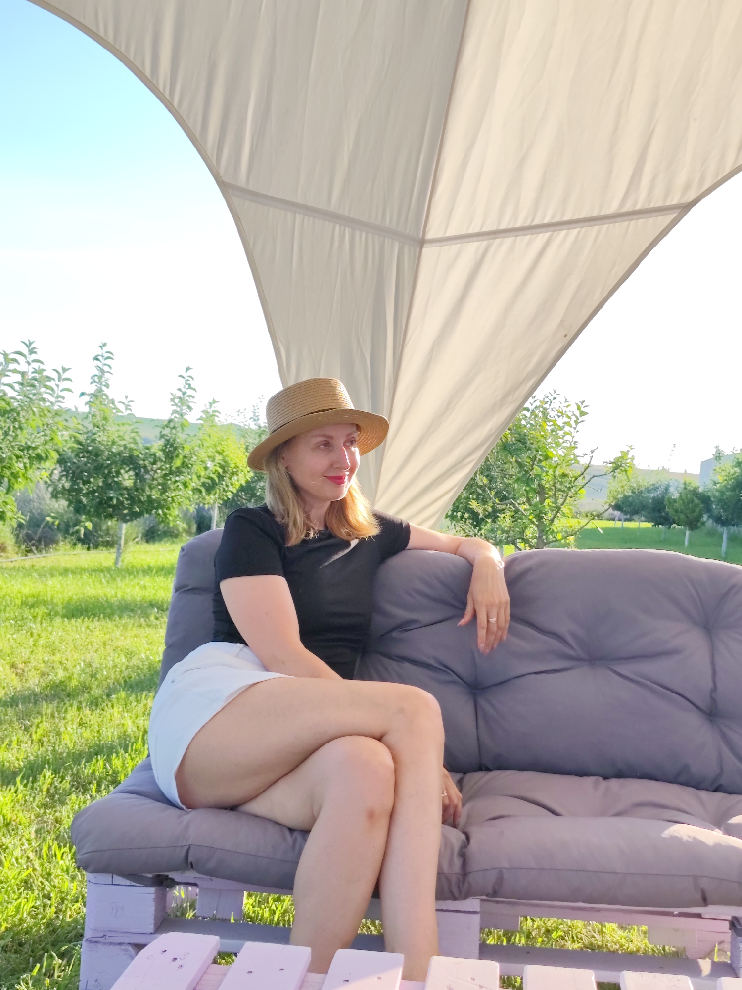 A woman sits on a gray outdoor sofa, her legs crossed, under a large, light-colored canopy. She is wearing a black t-shirt and white shorts, and a straw hat. Her head is turned to her right, and she is looking into the distance with a slight smile. Her hair is blonde and falls just past her shoulders. She has a ring on her right ring finger.
The setting appears to be a garden or orchard, with rows of green trees in the background. The grass is lush and green, and the sky is a clear blue. The sun is shining brightly, suggesting it is daytime, likely late afternoon given the warm light. The overall mood is relaxed and peaceful. The sofa is made of grey cushions and rests on a base constructed from painted wooden pallets, which are a pale pinkish-purple color. The woman is the sole subject in the image. The location is Nechaiane, Ukraine.