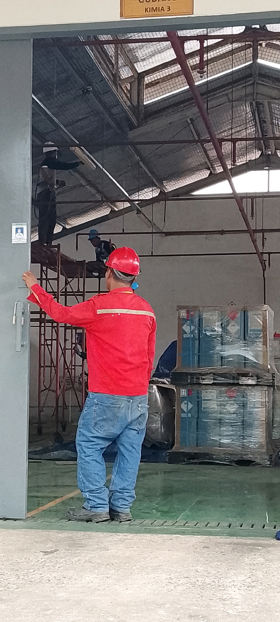 The image depicts the interior of a warehouse in Depok, Indonesia, specifically identified by a sign overhead that reads "GUDANG KIMIA 3" (Chemical Warehouse 3). In the foreground, a person wearing a red hard hat and a red long-sleeved work jacket with reflective stripes is facing away from the camera, looking towards the interior of the warehouse. They are also wearing blue jeans and dark work boots. Their right hand is extended, as if interacting with a grey mechanism on the left side of the frame.
Further inside the warehouse, two other individuals are visible. One, wearing a blue hard hat and a dark vest over a dark shirt, is working on scaffolding. Another person, mostly obscured by the scaffolding and the foreground subject, appears to be on the same scaffolding structure.
The warehouse environment is characterized by a corrugated metal roof with skylights, exposing wooden roof beams and red metal support structures. The floor is a green surface, with some areas appearing to be concrete. In the middle ground, stacked on wooden pallets and wrapped in clear plastic, are several blue containers, some with hazard symbols indicating they contain chemicals. These containers are positioned near the right side of the image. The overall scene suggests ongoing work or operations within a chemical storage facility.