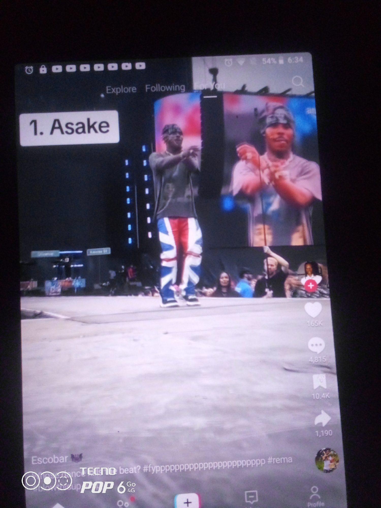 The image displays a screenshot of a mobile phone screen, likely from a social media platform like TikTok, showcasing a music performance video. The top of the screen shows standard phone interface elements including battery percentage (54%), time (6:34), and icons for signal strength and connectivity. Below this, navigation options like "Explore" and "Following" are visible.
The main content of the screen is a split view of a performance. On the left, a large white rectangle displays "1. Asake," indicating the artist or song at the top of a chart or ranking. To the right, two images of a performer wearing a bandana and a grey t-shirt are shown. The performer is captured mid-performance, with hands clasped, suggesting an energetic moment. They are wearing distinctive red, white, and blue striped pants, resembling a British flag design. In the background of the performance, a street scene with buildings and signs, one appearing to say "Kano St," is visible, suggesting an urban setting possibly in Nigeria, given the mention of "Howayi, Nigeria" in the prompt, though this location is not explicitly confirmed by visual cues within the image itself.
The lower portion of the screen reveals social media engagement metrics: "165K" likes, "4,815" comments, "10.4K" shares, and "1,190" saves. A profile picture of the performer is also displayed, along with various icons for interaction like a heart for likes and a comment bubble. At the very bottom, a caption is partially visible, containing "Escobar," "TECNO beat? #fyyyyyyyyyyyyyyyyyyyyyy #rema," and a TECNO POP 6 4G logo, further confirming the mobile device and likely its origin. The overall impression is a snapshot of a digital media experience focused on music and a performing artist.