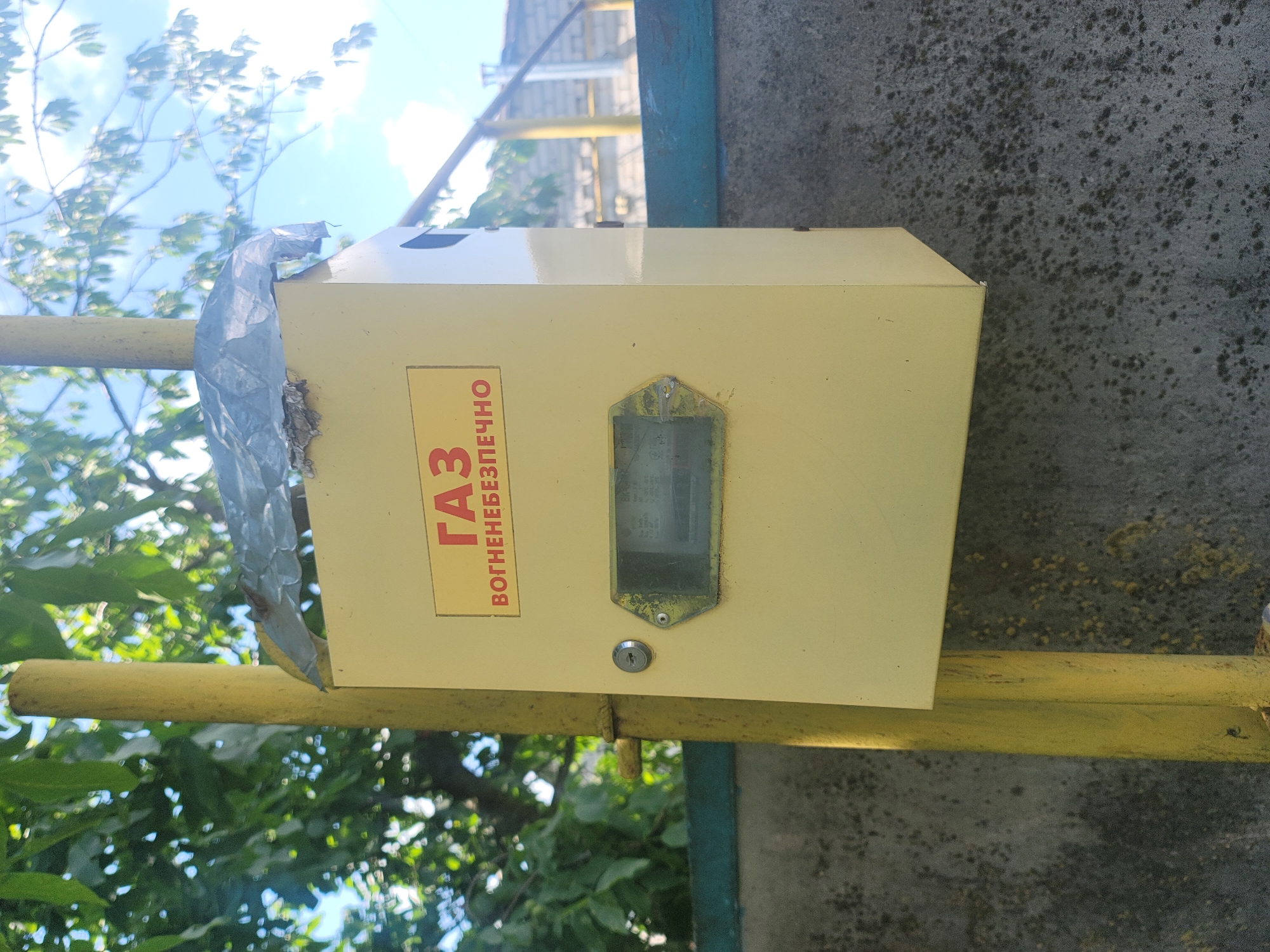 A yellow metal gas cabinet, approximately 30cm x 30cm x 20cm, is mounted on a yellow metal pole. The cabinet has a rectangular glass window displaying a gas meter. Above the window, a red and yellow sign reads "ГАЗ" and "ВОГНЕНЕБЕЗПЕЧНО," which translates to "GAS" and "FIRE HAZARD." A small keyhole is visible to the left of the window. The cabinet is slightly weathered and shows some signs of rust. The pole is also painted yellow and appears to be a gas line. In the background, there is a gray concrete wall, a blue painted fence, and green foliage from trees. The sky is visible in the upper portion of the image. The scene is likely located outdoors in a residential area in Vovkivka, Ukraine, given the cyrillic text on the sign and the typical appearance of such infrastructure in Eastern European countries.