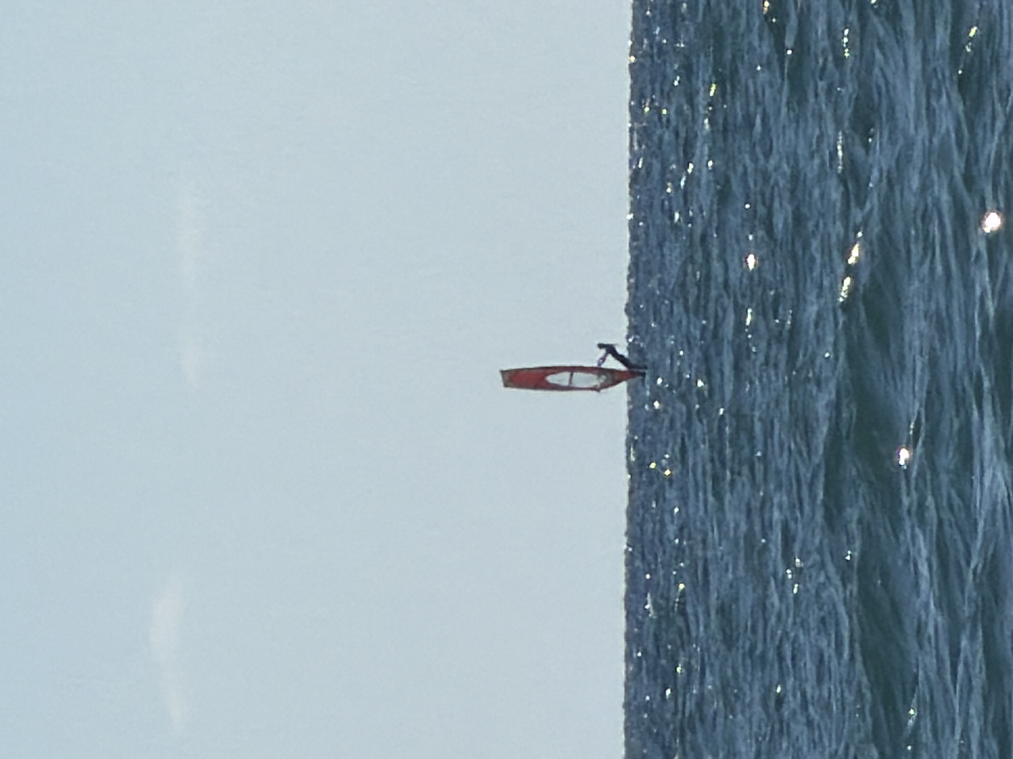 A lone windsurfer is seen from the chest up, with the sail obscuring the rest of their body. The windsurfer is positioned in the middle of the frame, in the sea, with a clear sky above. The sea is a dark blue, with small whitecaps and the sun glinting off the water. The sky is a light blue with a few wispy white clouds. The windsurfer's sail is red and white and is tilted to the right. The windsurfer appears to be moving to the right, away from the viewer. The image captures a serene and peaceful moment of recreation on the water.