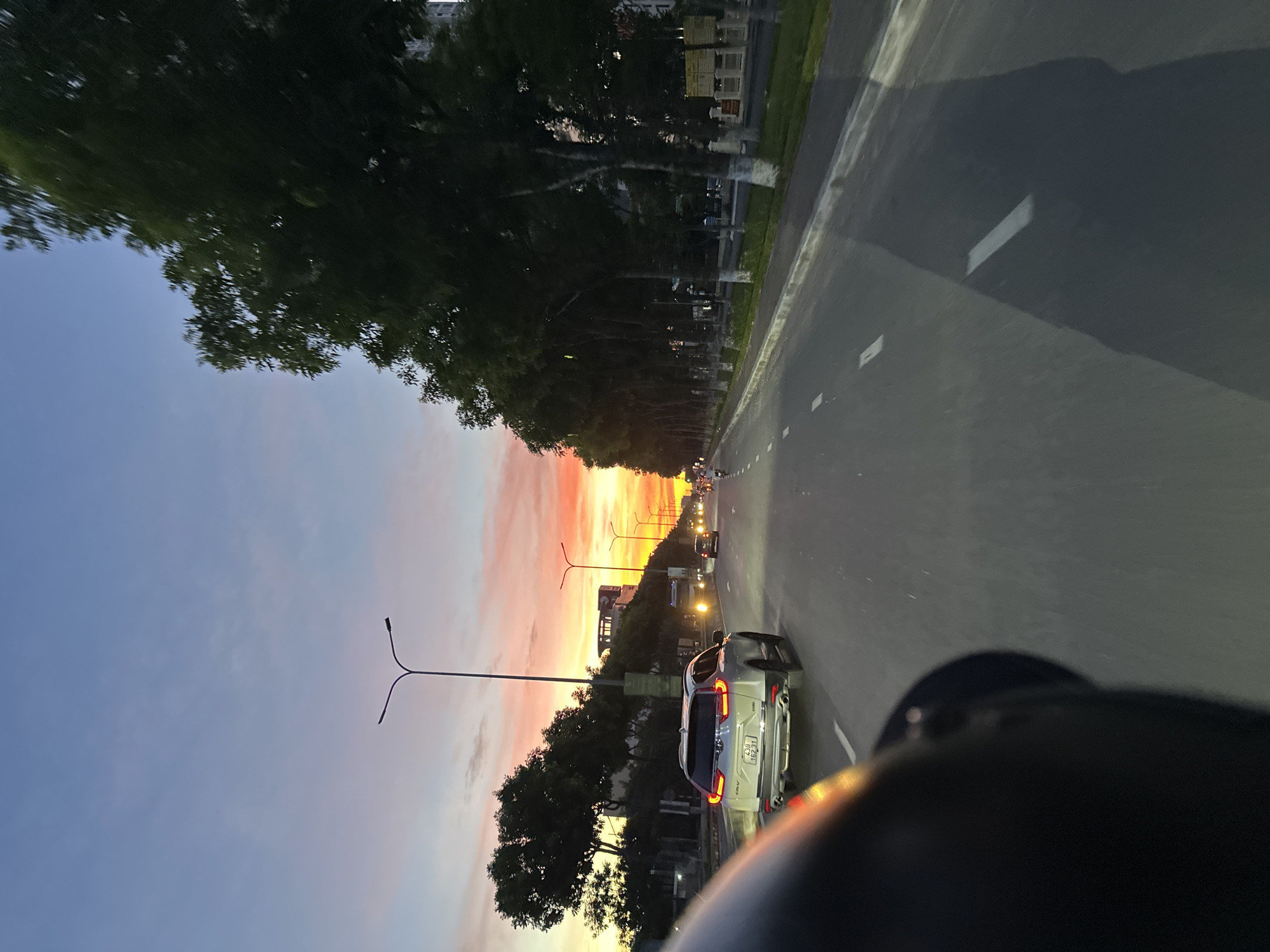 The image depicts a street scene in Đà Nẵng, Vietnam at sunset. A silver SUV is driving on the right side of the road, its taillights illuminated. The sky displays a gradient of colors, transitioning from deep blue at the top to vibrant orange and pink near the horizon, indicating the sun is setting. Tall trees line the right side of the road, casting shadows onto the asphalt. Streetlights are visible, some illuminated, along the roadside. The perspective suggests the photo was taken from a moving vehicle, possibly a motorcycle, as indicated by the blurred dark object in the foreground. Other vehicles and motorcycles are present in the distance, suggesting moderate traffic. Buildings and urban infrastructure are visible in the background. The overall atmosphere is one of a typical urban evening commute in a coastal city.