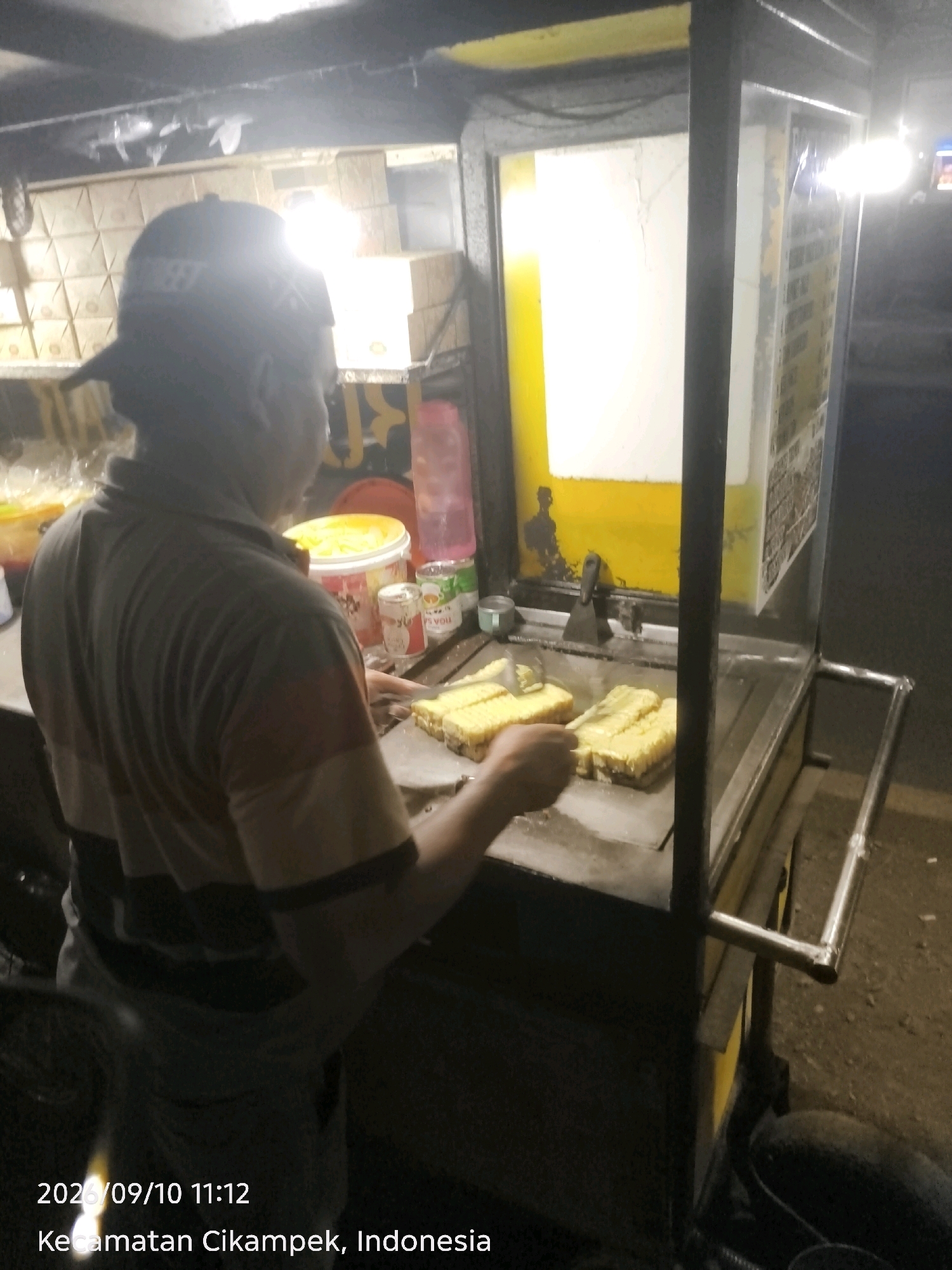 The image shows a man preparing food at an outdoor food stall in Pagaden, Indonesia. He is wearing a cap and a t-shirt, and is seen working on a flat-top grill. On the grill, there are several pieces of what appear to be bread, some of which are being spread with a creamy substance. The food stall has a bright yellow background and metal framing. Various containers and ingredients are visible on a shelf behind the grill. The lighting suggests it is either dusk or nighttime. The timestamp and location overlay at the bottom of the image read "2026/09/10 11:12 Kecamatan Cikampek, Indonesia".