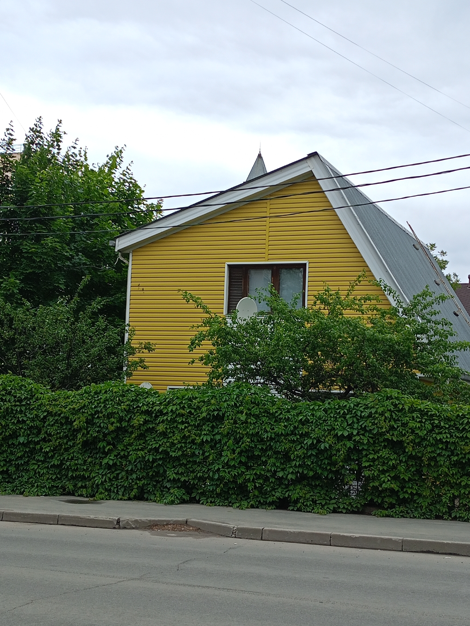 A bright yellow, two-story house with a steeply pitched gray metal roof stands behind a dense green hedge. The house has white trim around the roofline and a single window visible on the second story, which appears to be a double-hung window with dark brown shutters. A small satellite dish is mounted on the exterior wall to the left of the window, and another, larger satellite dish is visible just below the window. The house is surrounded by lush green trees and foliage, with a thick, leafy hedge forming a barrier in front of it.
In the foreground, a gray asphalt road runs parallel to a concrete sidewalk that borders the hedge. The sidewalk appears to be slightly damaged in places, with small potholes and debris. The sky above is overcast, suggesting a cloudy or possibly rainy day. The scene is framed by power lines running across the top of the image. There is no visible text in the image. The overall impression is of a residential area on an overcast day, with the vibrant yellow of the house contrasting with the surrounding greenery.