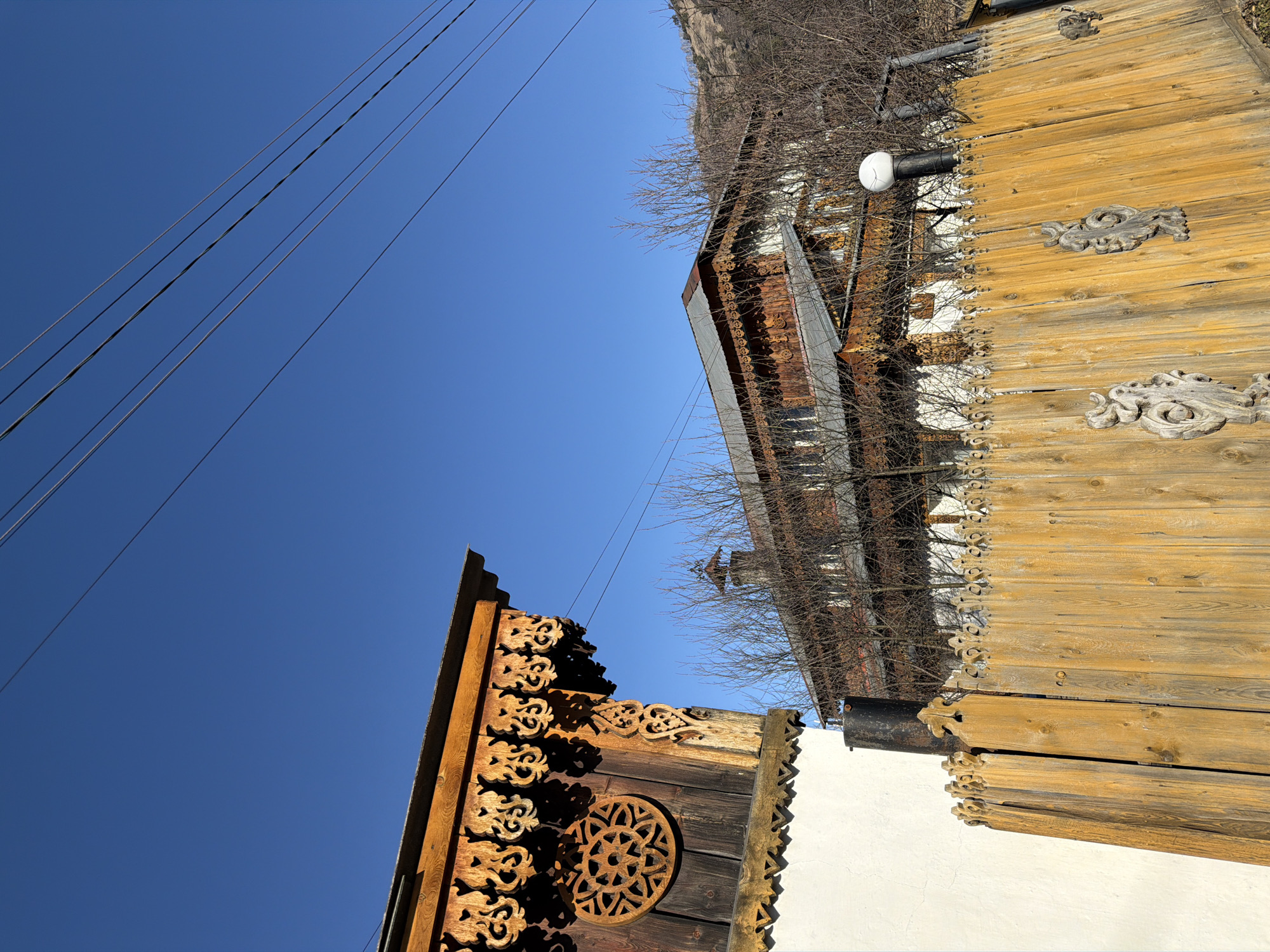 The image presents a low-angle view of buildings in Manzherok, Russia, against a clear blue sky. On the left, a section of a wooden building features intricate carvings, including a decorative rosette. A set of power lines runs parallel to the left edge of the frame. To the right, another building exhibits rustic wooden siding and additional ornate carvings along its edge. Bare tree branches intermingle with the buildings, suggesting it may be winter. A white, cylindrical light fixture is visible on the side of the right-hand building. The camera is angled upward, emphasizing the height of the structures.