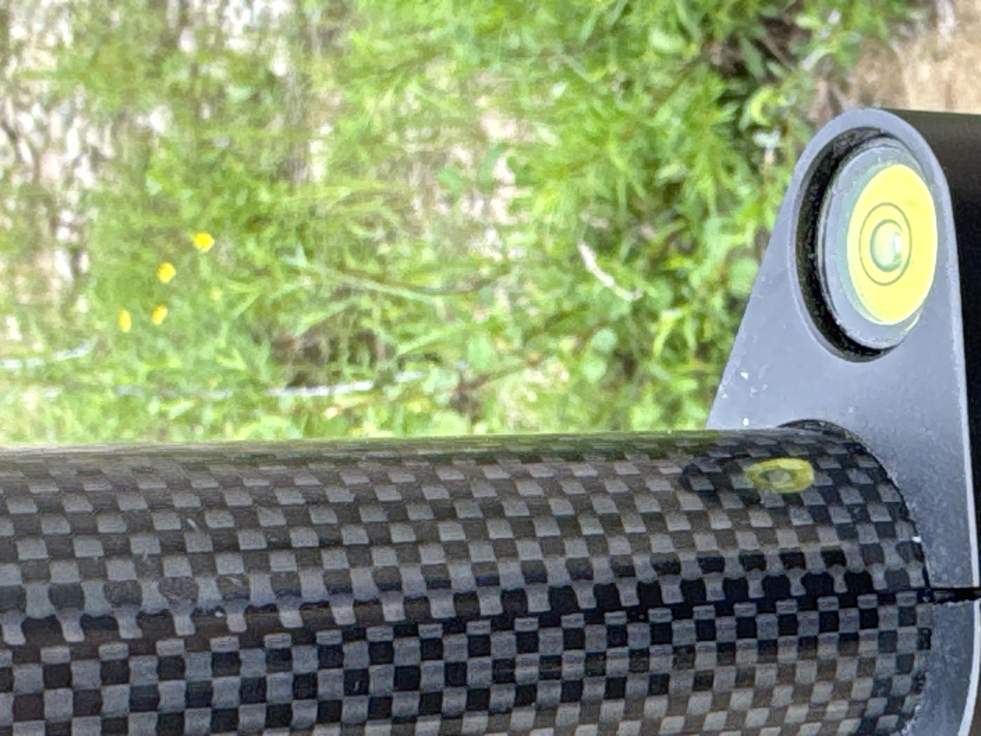 This close-up, eye-level shot focuses on a carbon fiber tube, likely part of a piece of equipment such as a camera tripod or a surveying tool. The tube has a distinctive checkered pattern of black and grey squares. To the right of the tube, a silver bracket is visible, housing a circular spirit level with a yellow bubble. The bubble is positioned slightly off-center, indicating the surface is not perfectly level. In the background, out of focus, is a lush green environment with scattered small yellow flowers, suggesting an outdoor setting in a natural landscape. The lighting appears to be natural daylight, possibly mid-morning or afternoon, and the weather seems pleasant and clear. The scene evokes a sense of fieldwork or outdoor activity, possibly related to photography, construction, or scientific measurement. No text is visible in the image. The location is identified as Murtygit, Russia.