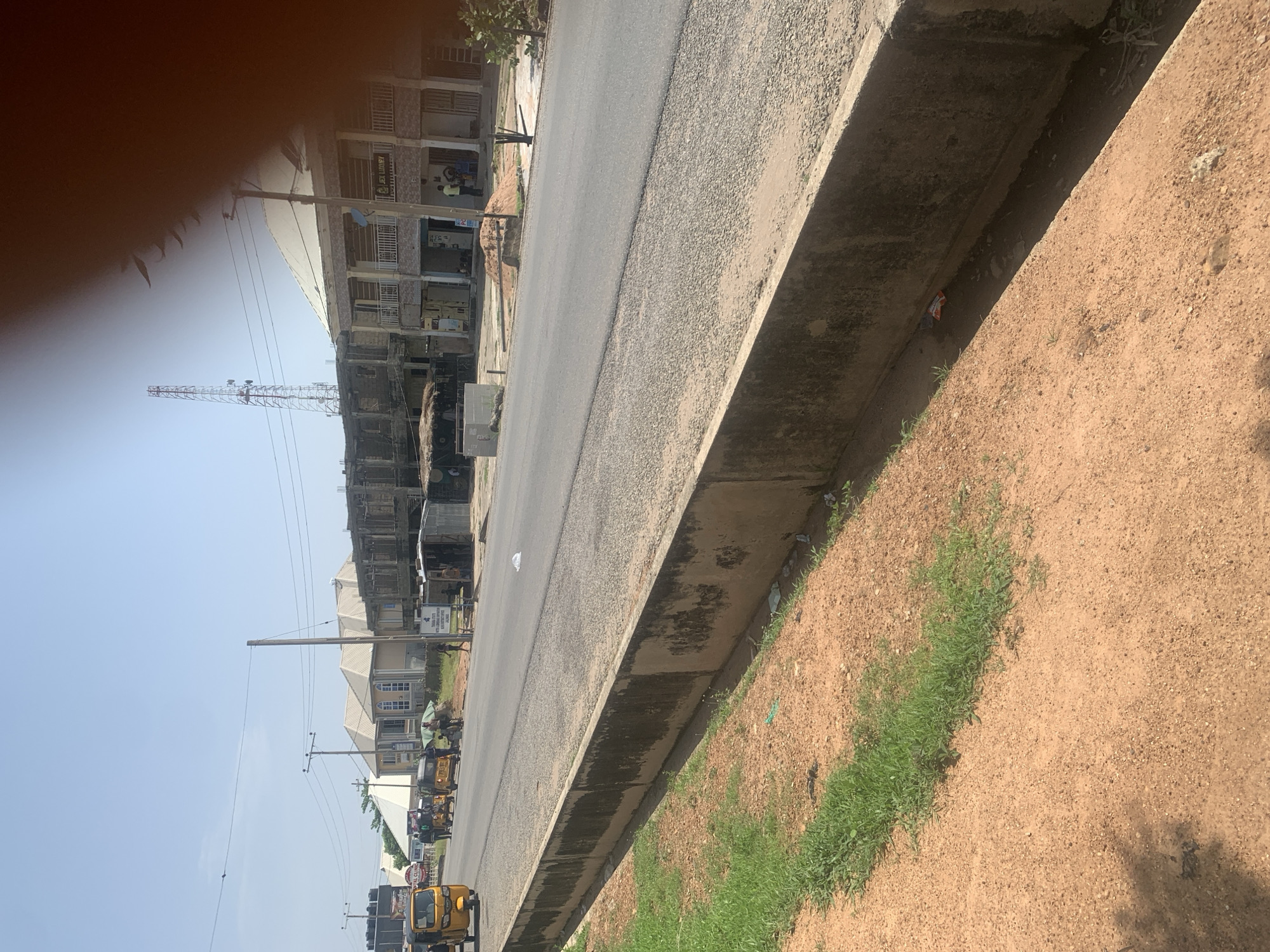 The image captures a street scene in Jalingo, Nigeria, likely during the daytime under a clear sky. In the foreground, a concrete drainage channel runs along the edge of a dirt and gravel road. Sparse green grass grows along the inner edge of the drainage. The road itself is paved with asphalt and appears to be moderately traveled.
Several yellow auto-rickshaws, known as "Keke Napep" or "Marwa," are visible on the road. Some are stationary, while one is in motion. A few individuals are present near the vehicles, with some appearing to be passengers or operators.
Buildings line the opposite side of the road, displaying a mix of architectural styles. One building has a prominent "JEX LUXURY" sign. Construction appears to be ongoing or recent on some of the structures, with visible scaffolding and unfinished sections. A tall telecommunications tower stands in the background, indicating modern infrastructure in the area. The overall impression is of a typical urban environment in Jalingo, with active street life and ongoing development.