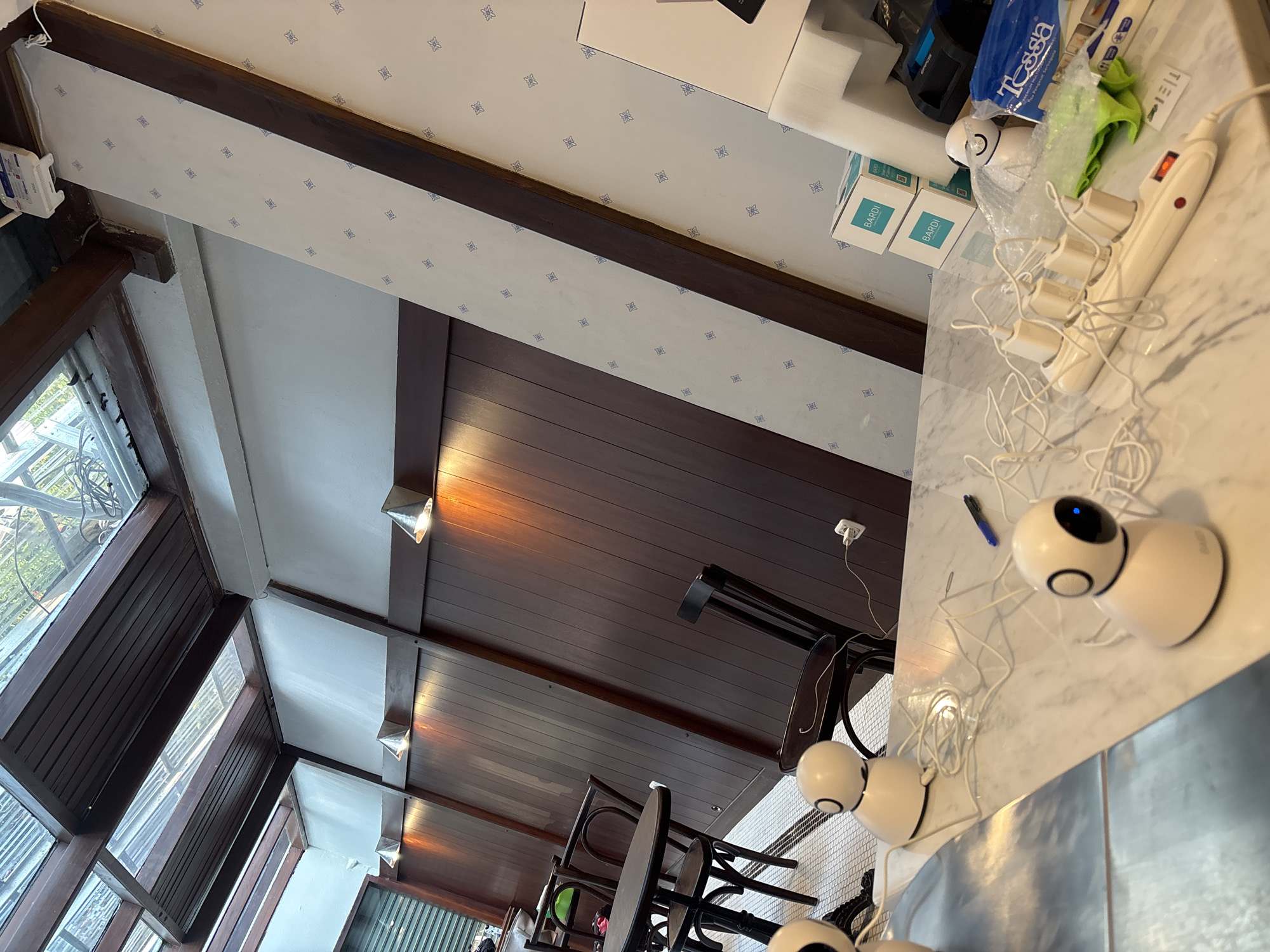 The image depicts the interior of what appears to be a cafe or restaurant in South Jakarta, Indonesia. The scene is illuminated by natural light filtering through a sloped glass ceiling and supplemented by spotlights mounted on wooden panels. The walls are a combination of dark wood paneling and patterned wallpaper.
In the foreground, a marble countertop is visible with several white, egg-shaped electronic devices, possibly smart cameras, connected by white cords to a power strip. One device has a blue light indicating it is active. A black pen lies on the countertop next to the devices.
Further into the background, several dark wooden chairs are arranged around a dark circular table. Another dark wooden chair is positioned against the wall. The floor is tiled with a black and white mosaic pattern.
On the right side of the frame, stacked cardboard boxes with "BARDI" branding are visible, along with other electronic equipment.
The overall atmosphere suggests a modern, indoor commercial space, possibly with a focus on technology, given the presence of multiple electronic devices.