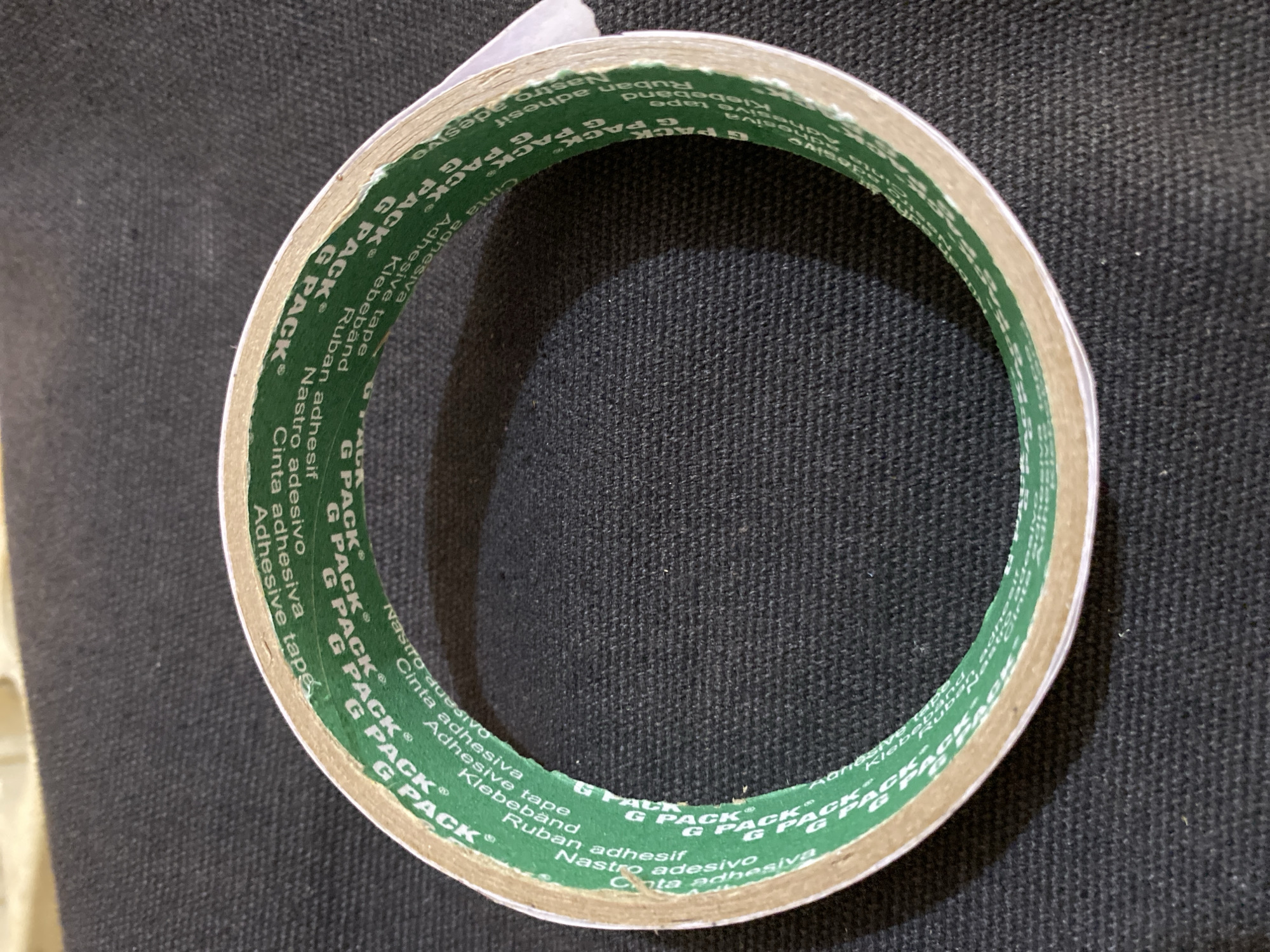 A roll of green double-sided adhesive tape with white lettering is centered on a dark, textured surface, likely a fabric. The tape is circular and appears to be unwound, with a small portion of the white liner peeking out from the edge. The lettering on the tape repeats "G PACK" in white, along with other words in different languages, such as "adhesive tape," "Nastro adesivo," "Cinta adhesiva," and "Klebeband." The background surface is a consistent dark gray with a visible weave pattern, suggesting it could be upholstery, a dark textile, or a similar material. The lighting creates a soft shadow cast by the tape roll onto the surface below, indicating a light source from the top. The image quality is clear, showing the details of the tape's printing and the texture of the background. There are no people, animals, or any other objects present in the scene. The location in Lhokseumawe, Indonesia, is not depicted or suggested by any visual elements in the image itself.