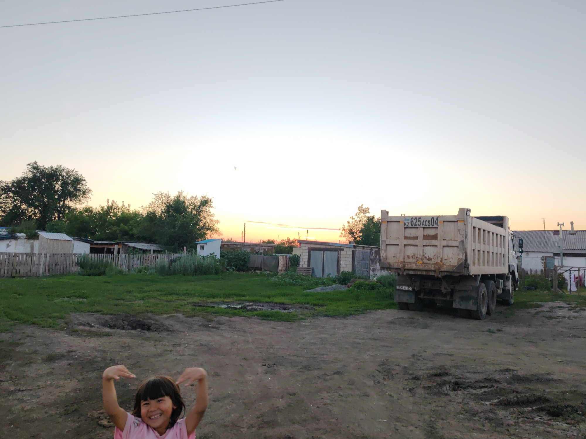 In this outdoor scene, a young girl with dark hair, wearing a pink t-shirt, is captured with her arms raised and hands open, smiling broadly at the camera. The setting appears to be a rural or semi-rural area, possibly a village in Akzhar, Kazakhstan, given the context. In the background, several small, weathered buildings with corrugated metal roofs and wooden fences are visible. A large, white dump truck with the license plate "kz 625 ACS 04" is parked to the right of the frame. The ground is predominantly dirt and patchy grass. The sky above is a pale gradient of blue and white, with the sun setting on the horizon, casting a soft light over the scene. The overall impression is a candid moment captured in a humble, outdoor environment.